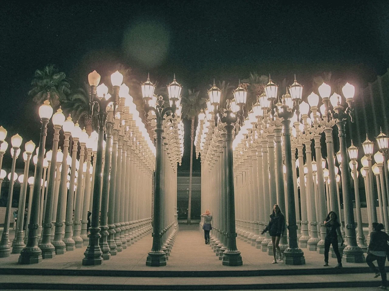 Night view of the Urban Light art installation with numerous street lamps arranged in a grid, with a few people walking and taking photos.