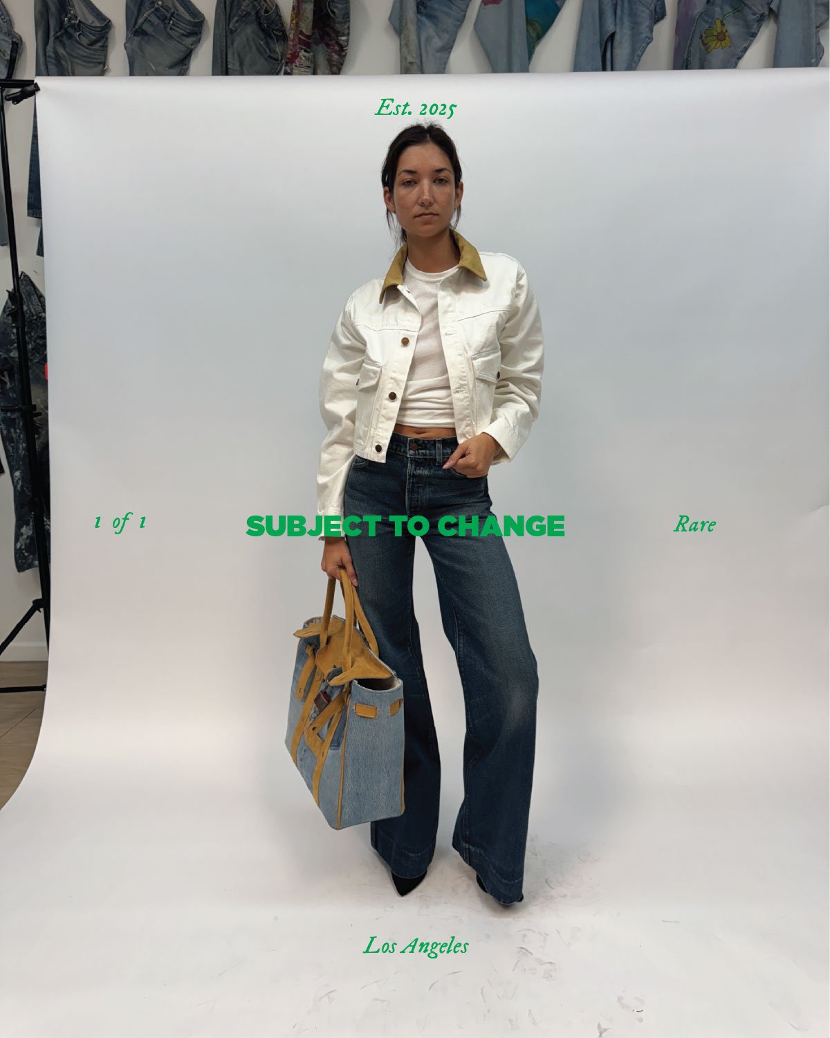 A woman standing in front of a white backdrop with green text that reads 'Est. 2025', '1 of 1', 'Rare', and 'Los Angeles'. She is wearing a white jacket with a tan collar, a white t-shirt, and dark jeans, holding a tan and blue handbag in her left ha