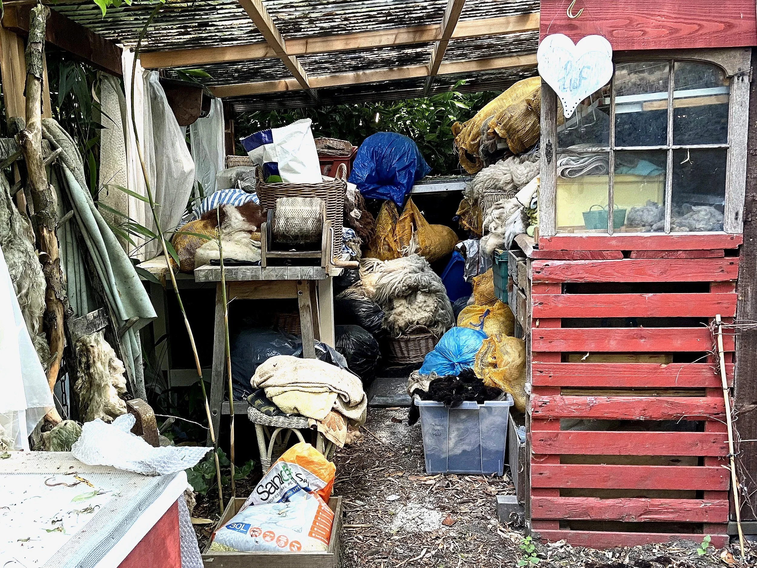 Ein Chaotischer Lagerraum mit verschiedenen Stoffen, Lumpen und Kleidung, umgeben von Holz- und Metallregalen, im Freien mit einem überdachten Dach