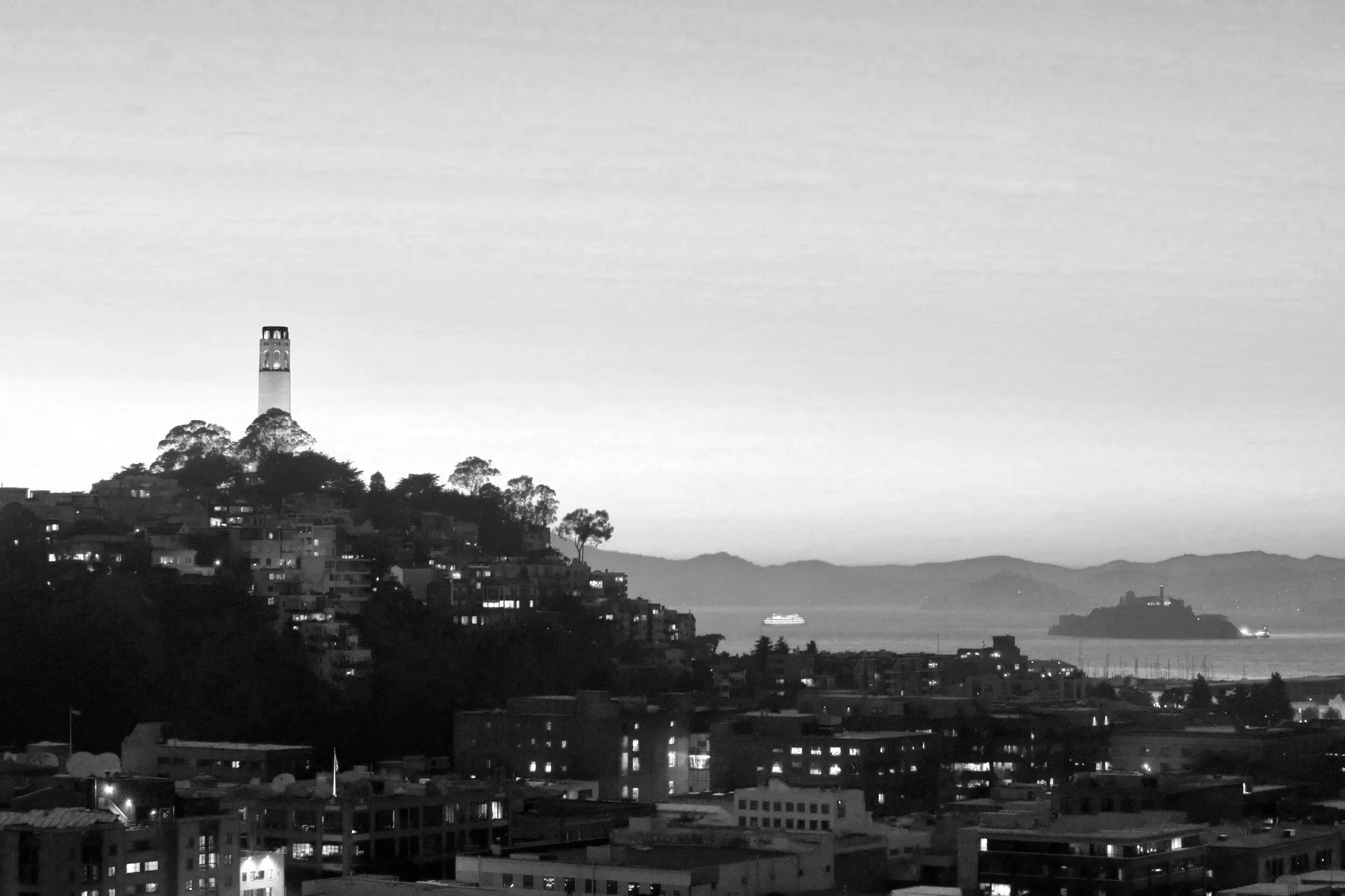 A black and white photo of a cityscape with a hill featuring the Coit Tower on top, overlooking a bay with boats and distant mountains in the background.