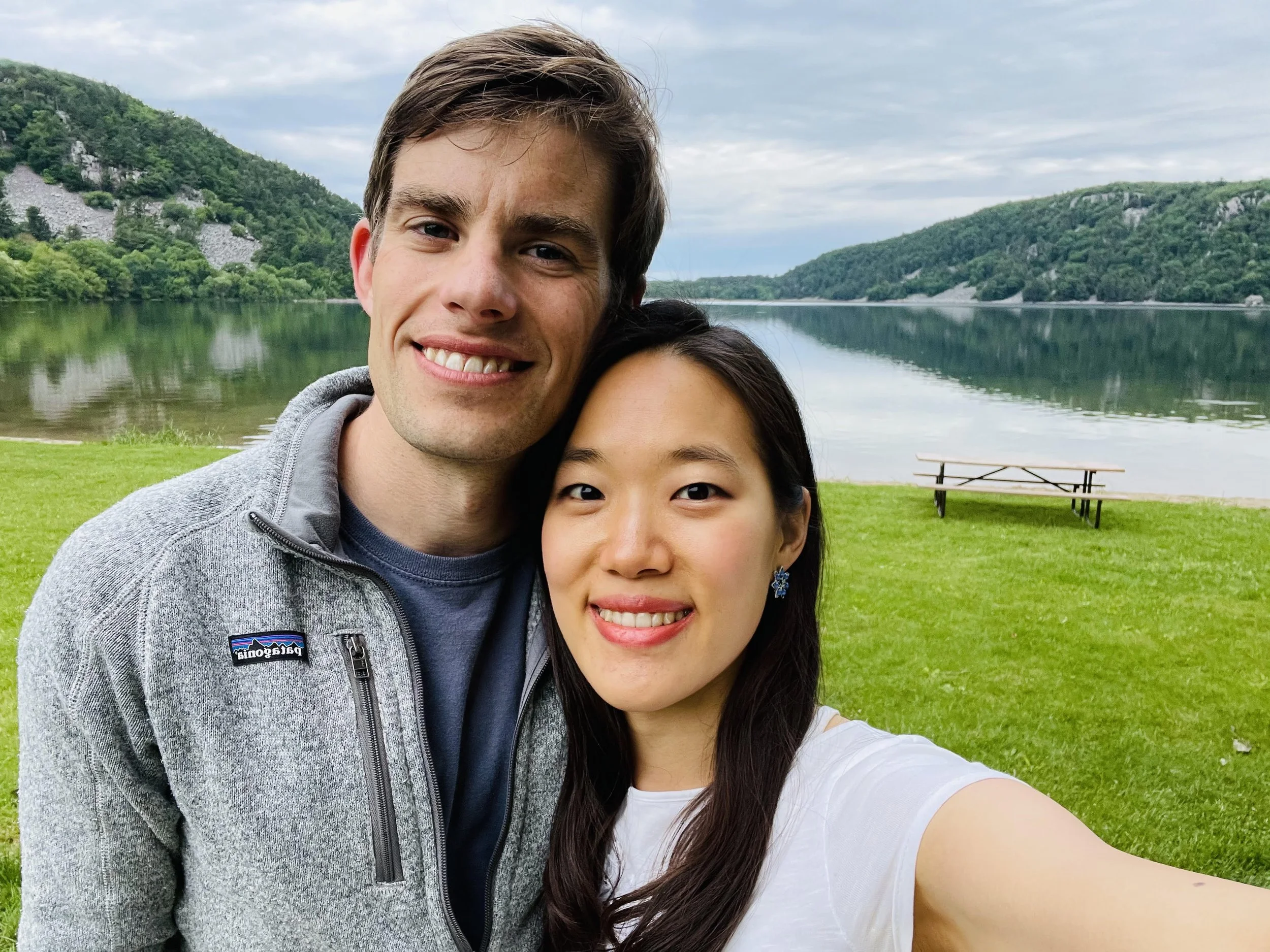 A young man and woman taking a selfie outdoors near a lake with hills in the background.