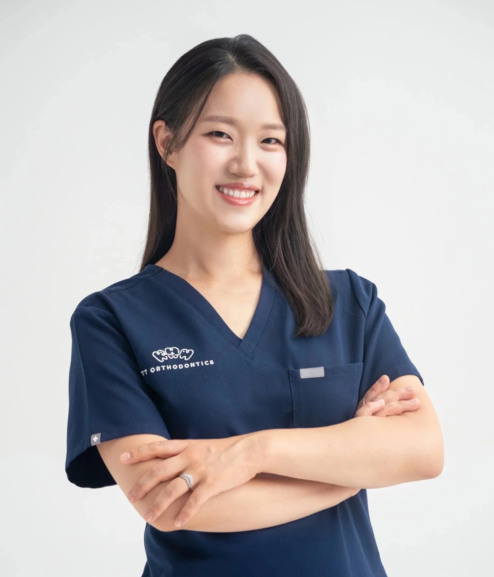 Smiling woman with dark hair wearing navy medical scrubs with teeth orthodontics logo, arms crossed against plain white background.