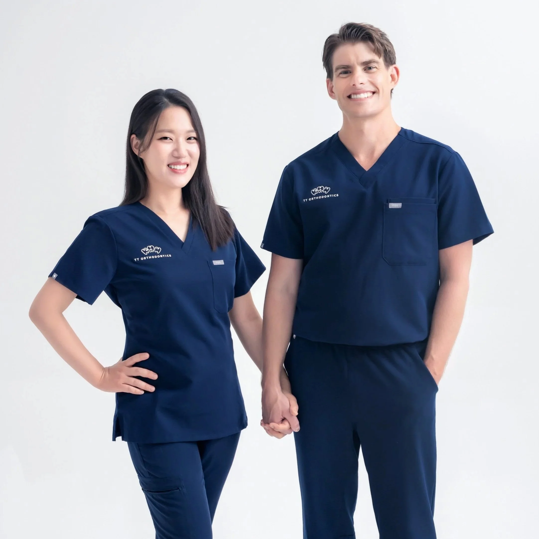 A man and a woman in matching navy scrubs holding hands and smiling, standing against a plain white background.