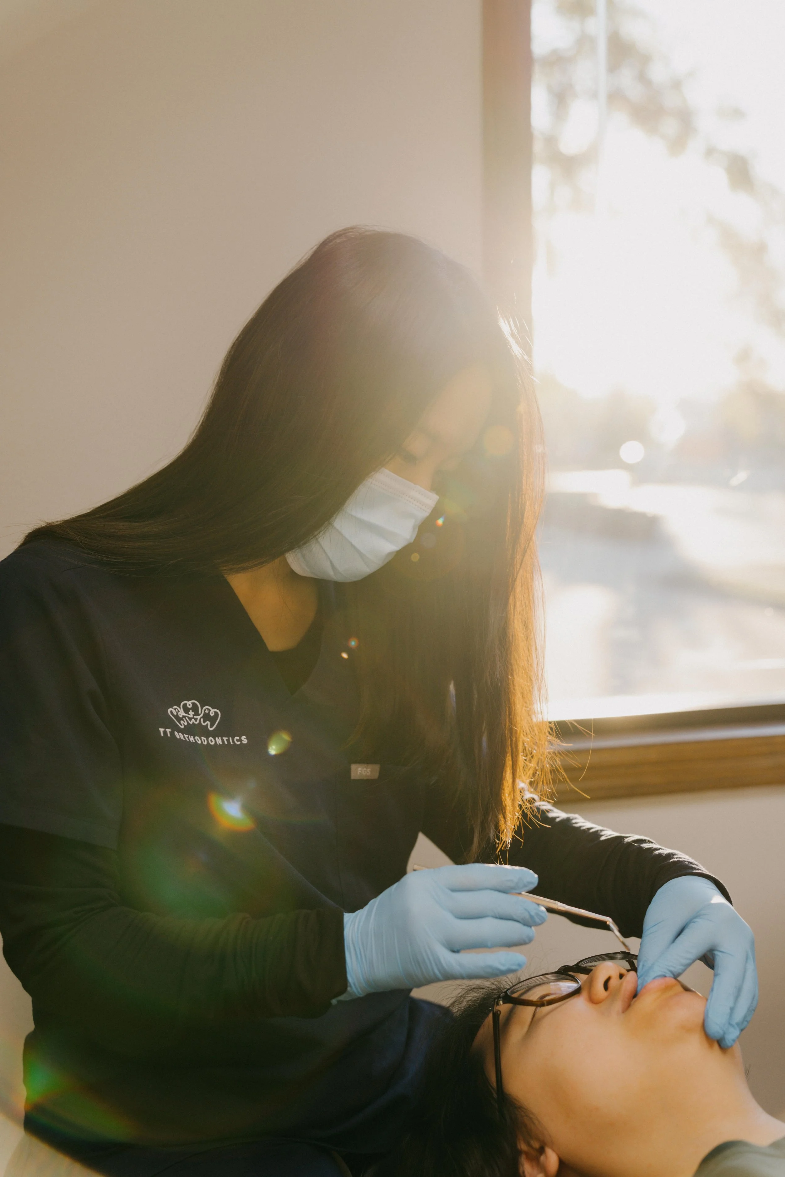Dr.Lee in scrubs and a face mask performs a medical procedure on another patient with glasses, lying on a bed, in a well-lit room with sunlight coming through a window.