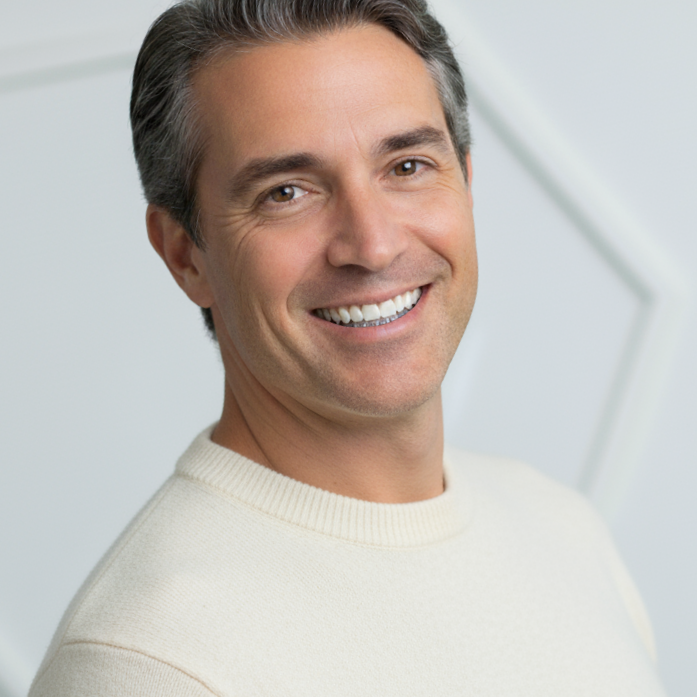 A smiling man with gray hair wearing a cream sweater in front of a neutral background.