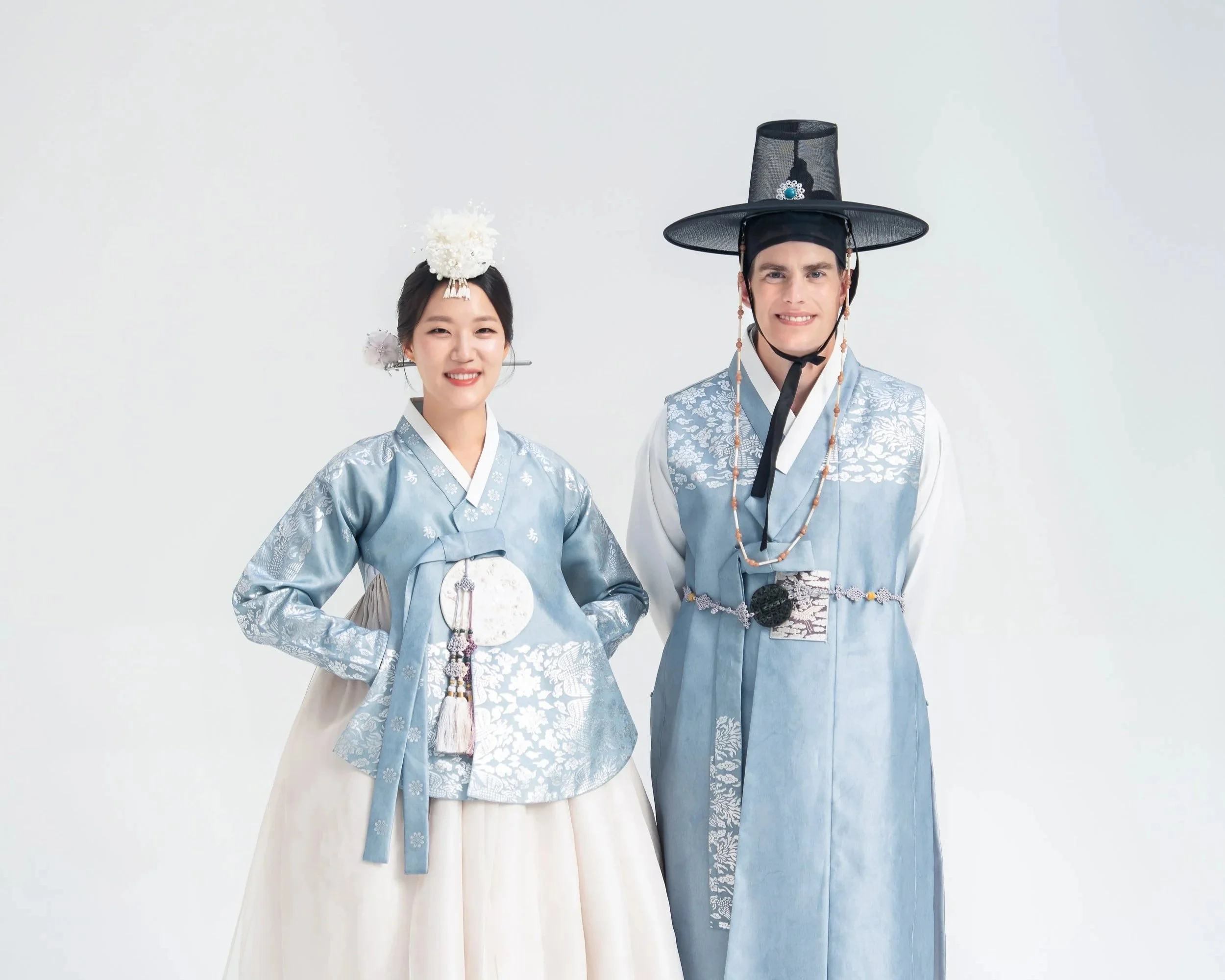 A man and woman in traditional Korean clothing, hanbok, standing side by side against a plain white background. The woman is wearing a light blue jeogori (top) with intricate designs, a white chima (skirt), and a decorative hairpiece. The man is dressed in a matching light blue garment with a hat called a gat.