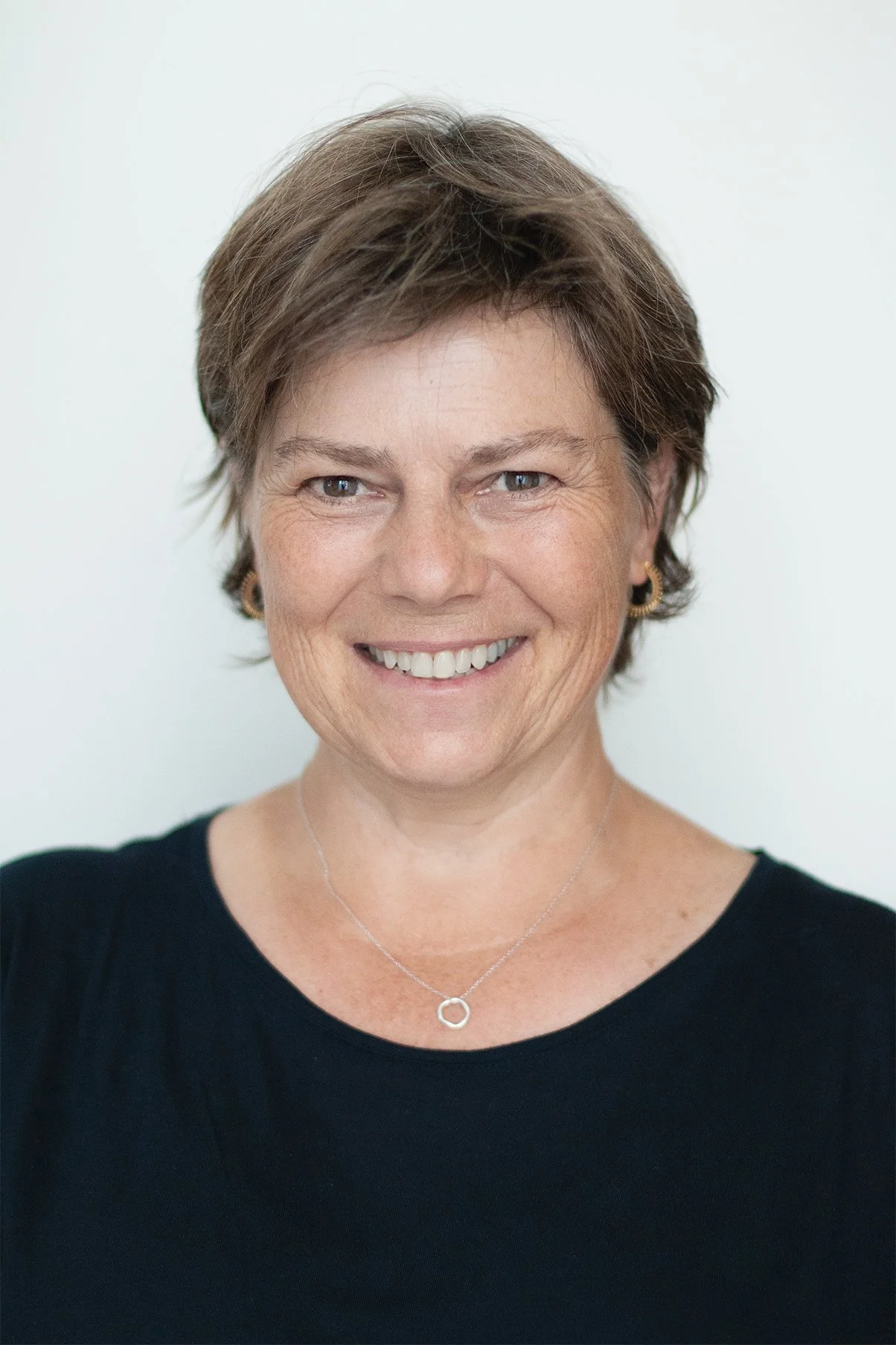 Porträt einer lächelnden Frau mit kurzen braunen Haaren, schwarzem Top vor einem weissen Hintergrund.