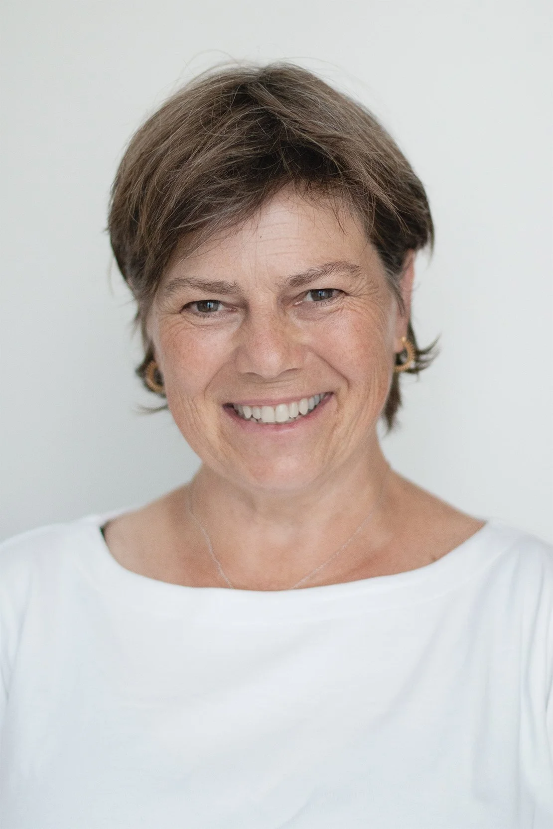 Porträt einer lächelnden Frau mit kurzen braunen Haaren und einem weissen Oberteil vor einem hellen Hintergrund.