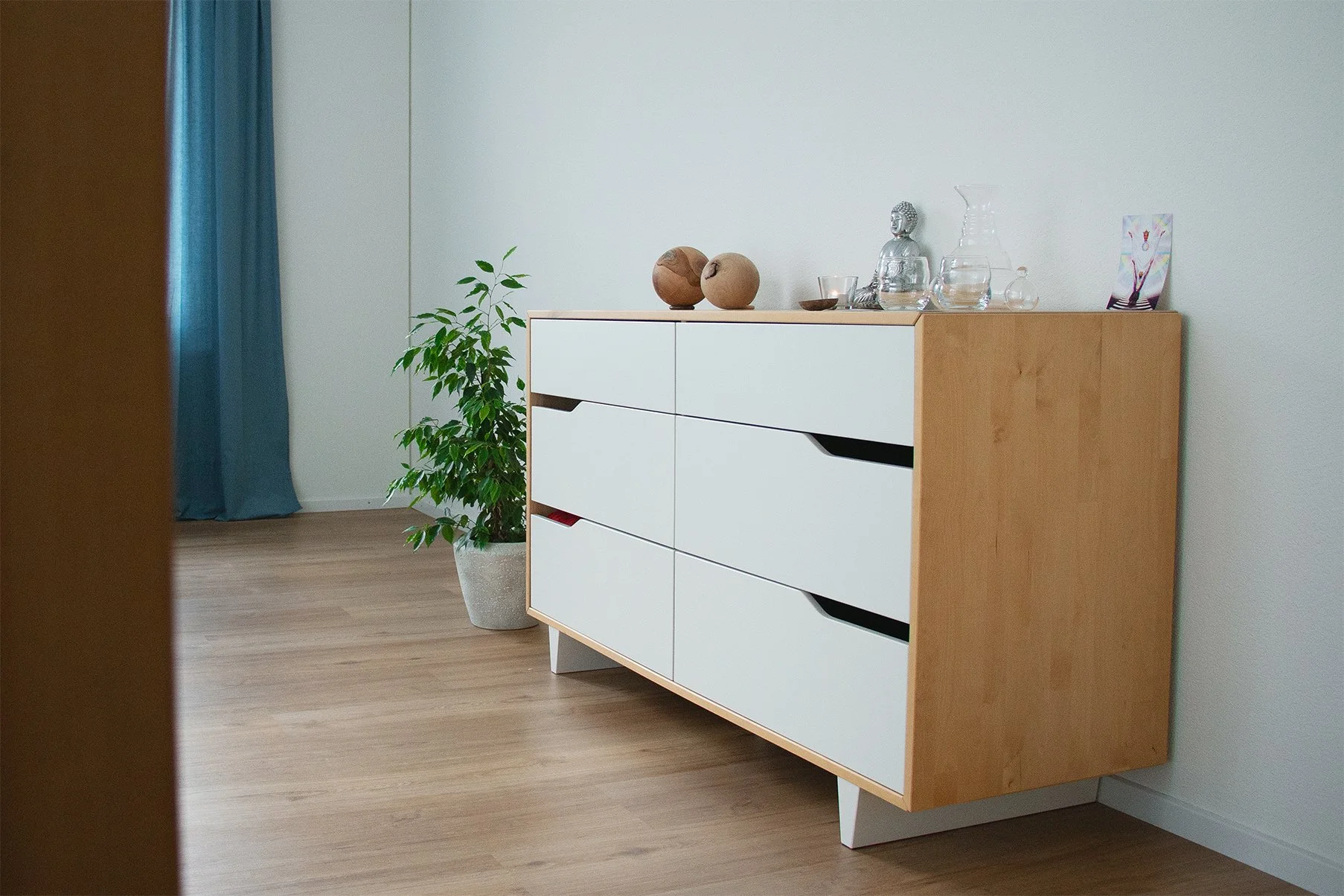 Moderner Kamin mit weißen Schubladen und natürlichem Holzrahmen, daneben eine Topfpflanze, Dekokugeln, Glaswaren und ein Farbpostkarten auf der Oberfläche, in einem Raum mit hellen Wänden und Holzboden.