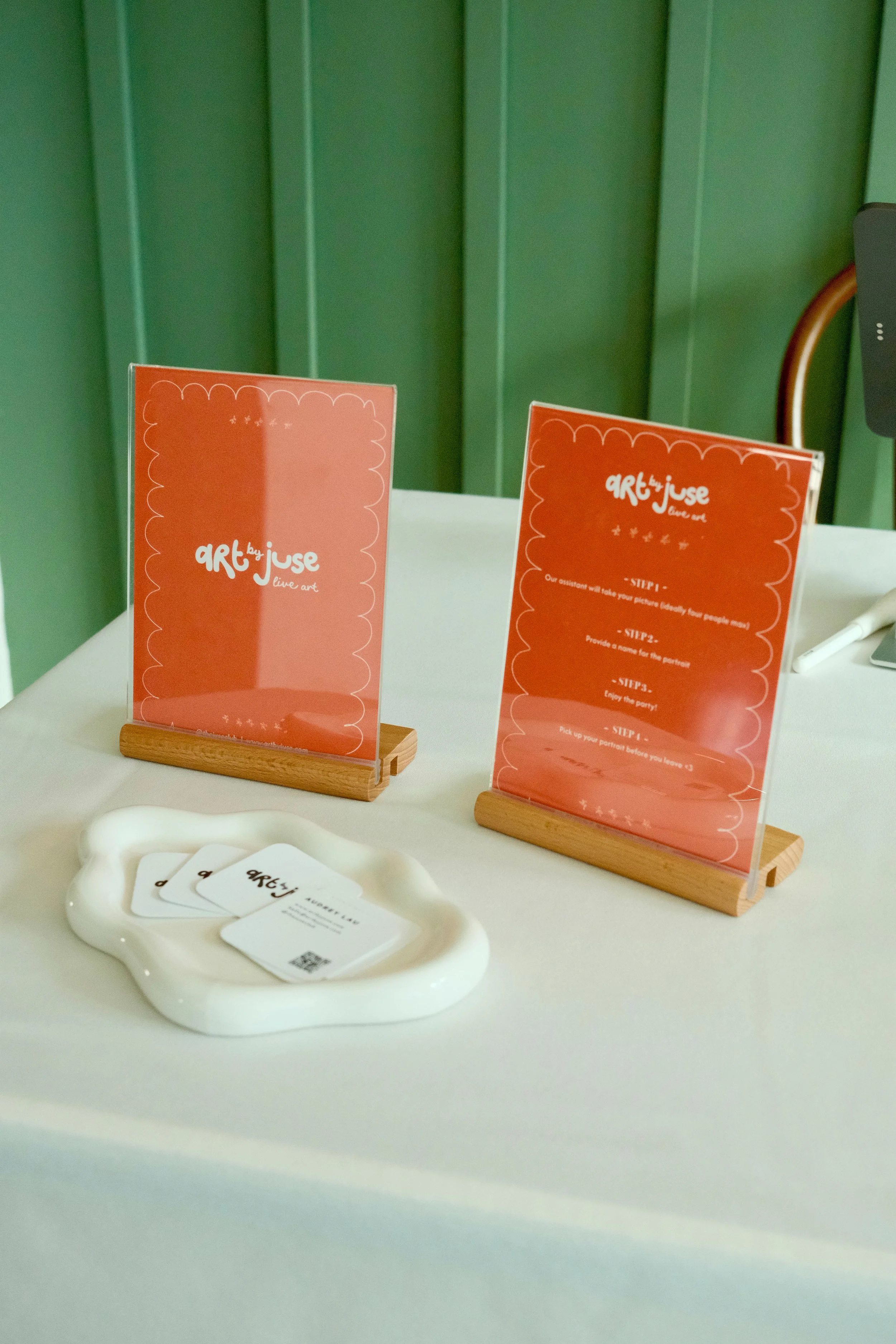 Table with two pink signs in wooden holders and a white dish holding business cards, with a green wall in the background.