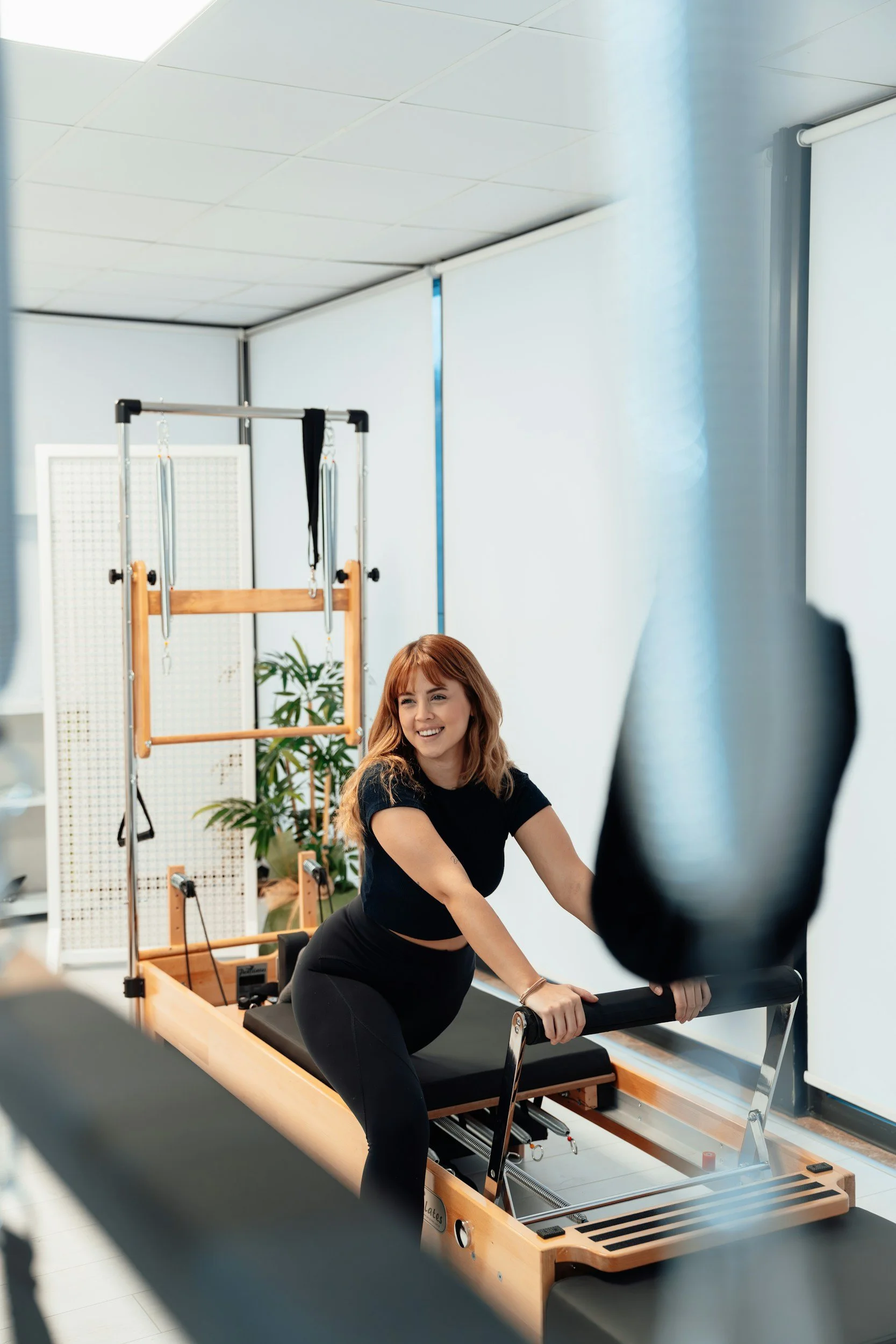 A woman in black workout clothes smiling while using a Pilates reformer machine in a fitness studio.