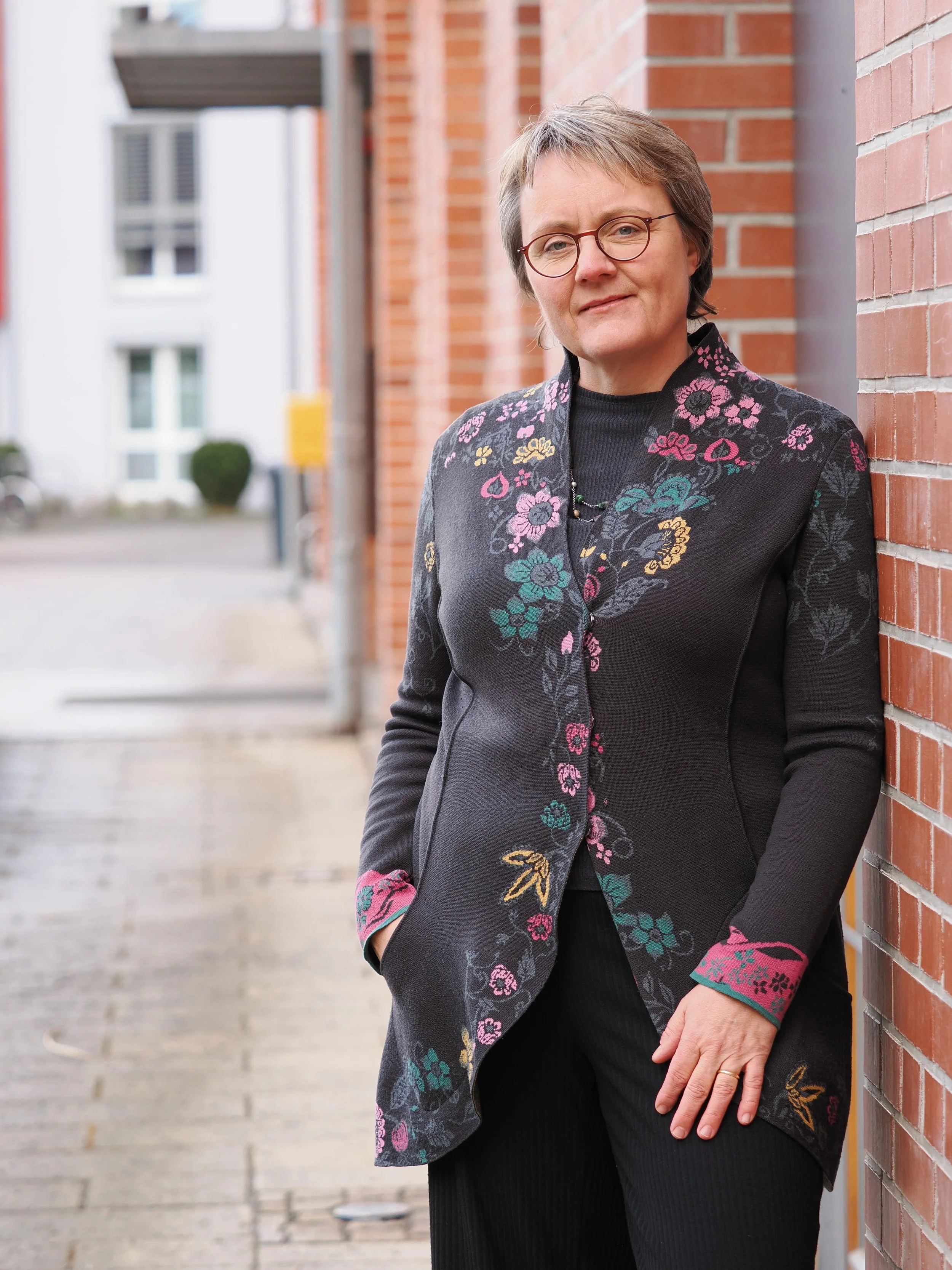 Fachjournalistin Katja Dorothea Buck lehnt an einer roten Backsteinmauer, im Hintergrund ein urbaner Straßenabschnitt mit Gebäuden und Büschen.