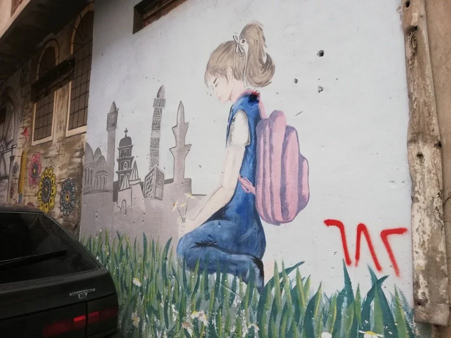 Streetkunst eines jungen Mädchens mit Rucksack, das auf Gras sitzt, im Hintergrund Silhouetten von Gebäuden und Stadtattrappen, Graffiti auf einer Wand.