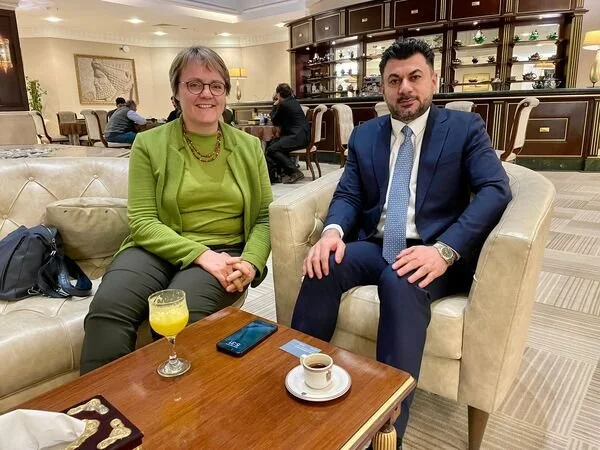Katja Dorothea Buck und ein Mann, sitzen in einem Hotel lobby oder einem Lounge-Bereich, trinken Kaffee und Saft, im Hintergrund Regal mit Dekorationsartikeln.