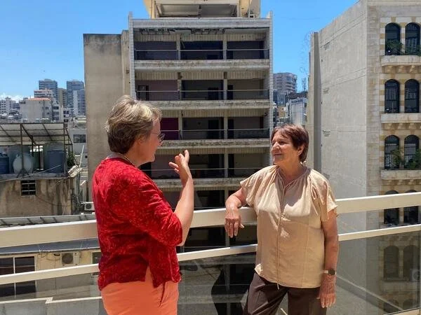 Zwei Frauen unterhalten sich auf einem Balkon in der Stadt, im Hintergrund liegen Hochhäuser.