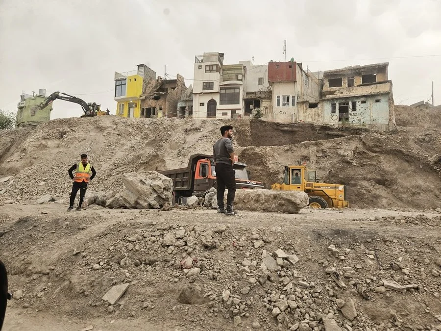Baustelle mit zwei Männern, Bagger und Traktor, vor zerstörten Häuser in Mossul