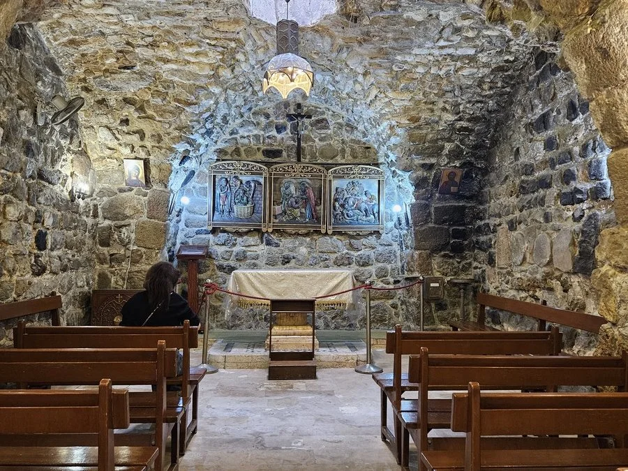 Kleine Kapelle mit Steinmauern, alte Gemälde und Ikonen, Holzbänke, eine Person sitzt im Vordergrund, Kerze und alter Kronleuchter.