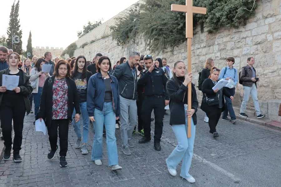 Christen in Palästina bei einer Prozession mit einer Frau, die ein Holzkreuz trägt, auf einer gepflasterten Straße neben einer Steinmauer.