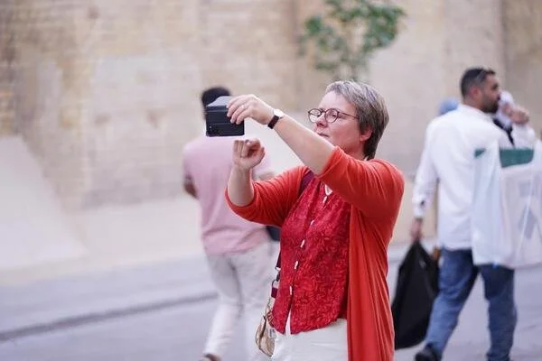 Eine Frau mit Brille und grauen Haaren, die ein Smartphone zum Fotografieren benutzt, auf der Straße. Im Hintergrund laufen Männer vorbei.