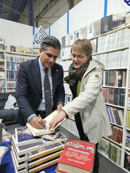 Ein Mann in Anzug und Katja Dorothea Buck schauen sich in einem Buchladen an. Sie blättern durch Bücher auf einem Tisch, umgeben von Bücherregalen.