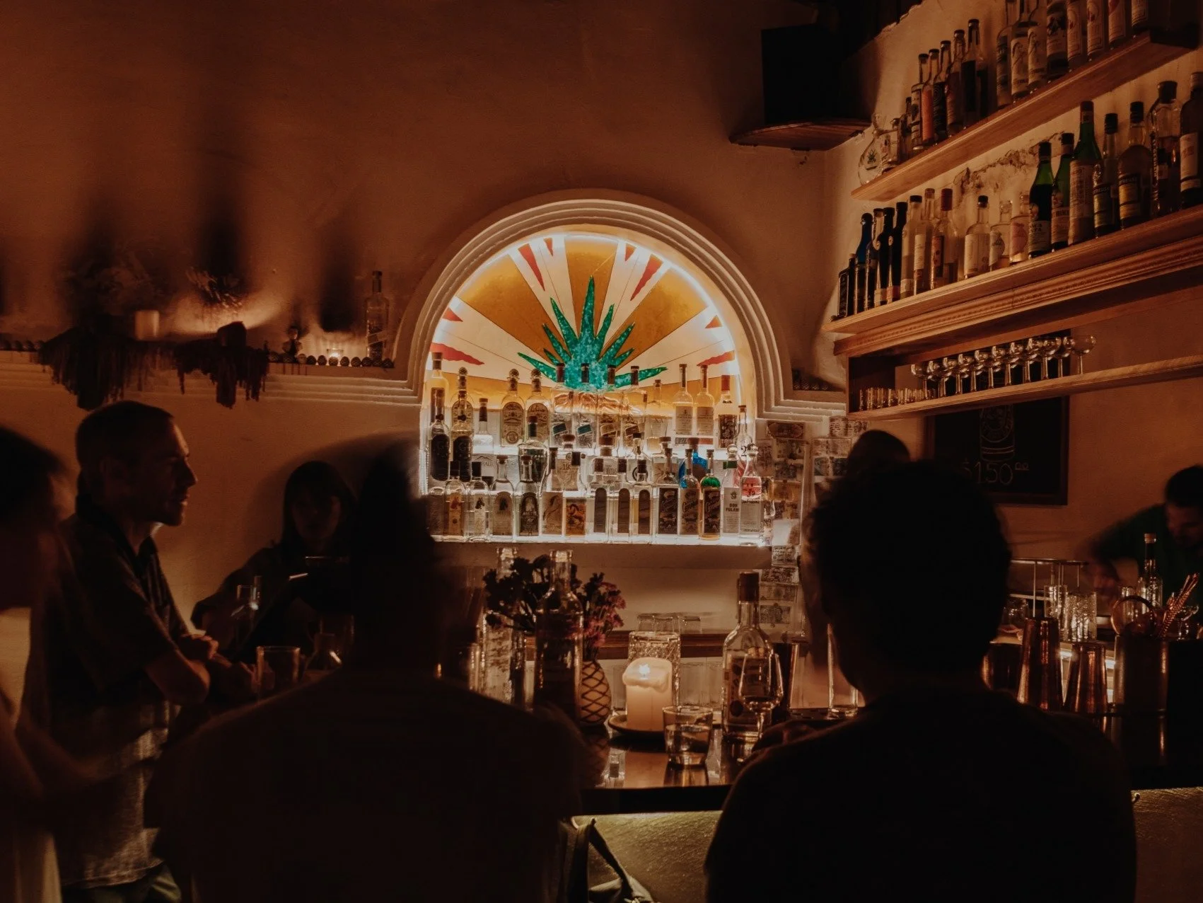 Interior de un bar con varias personas sentadas, estantes con botellas y una ventana con decoración en forma de sol y una planta en el centro.