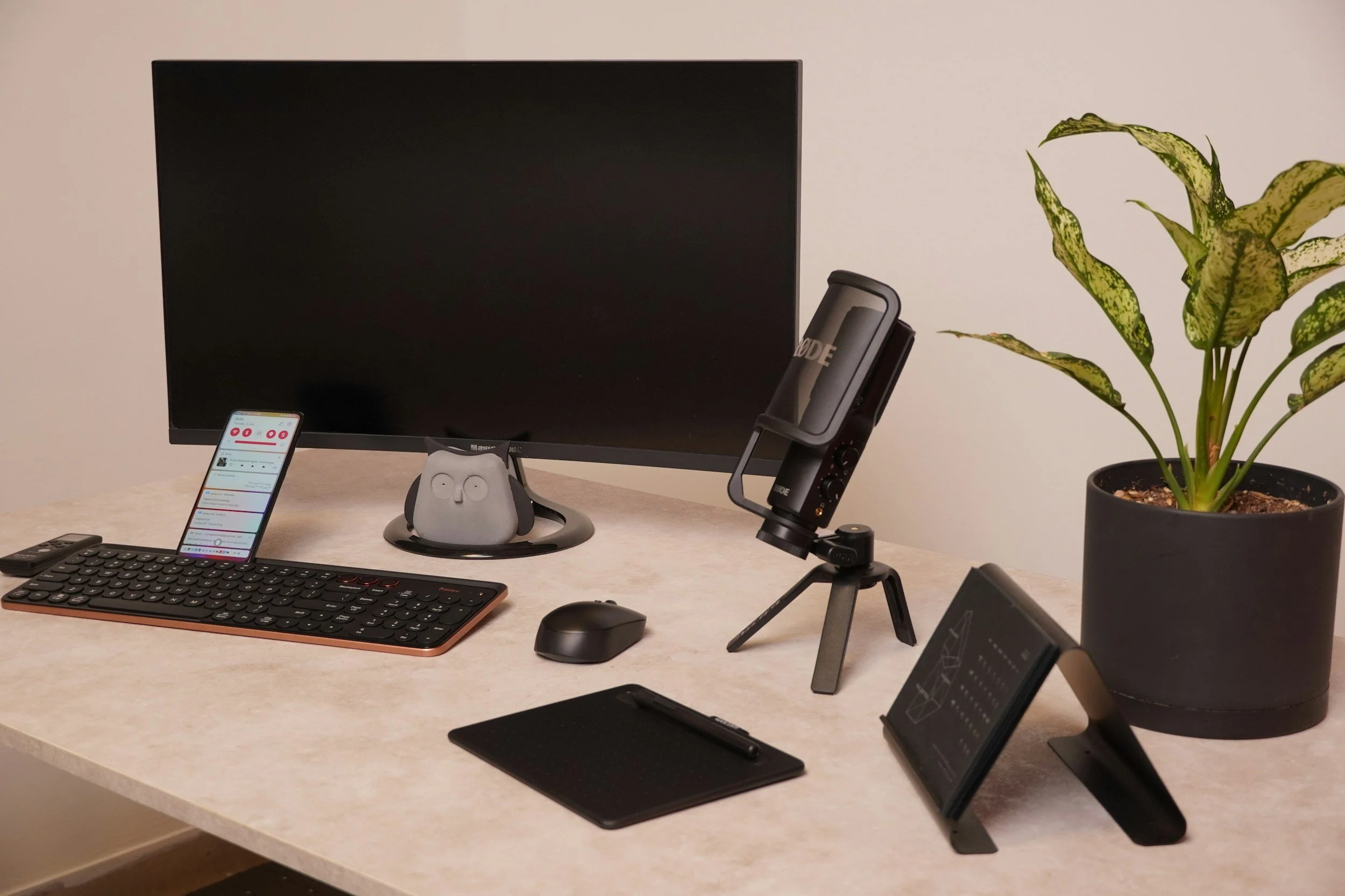 Home office desk with a computer monitor, smartphone, wireless keyboard, mouse, microphone, tablet, and a potted plant.