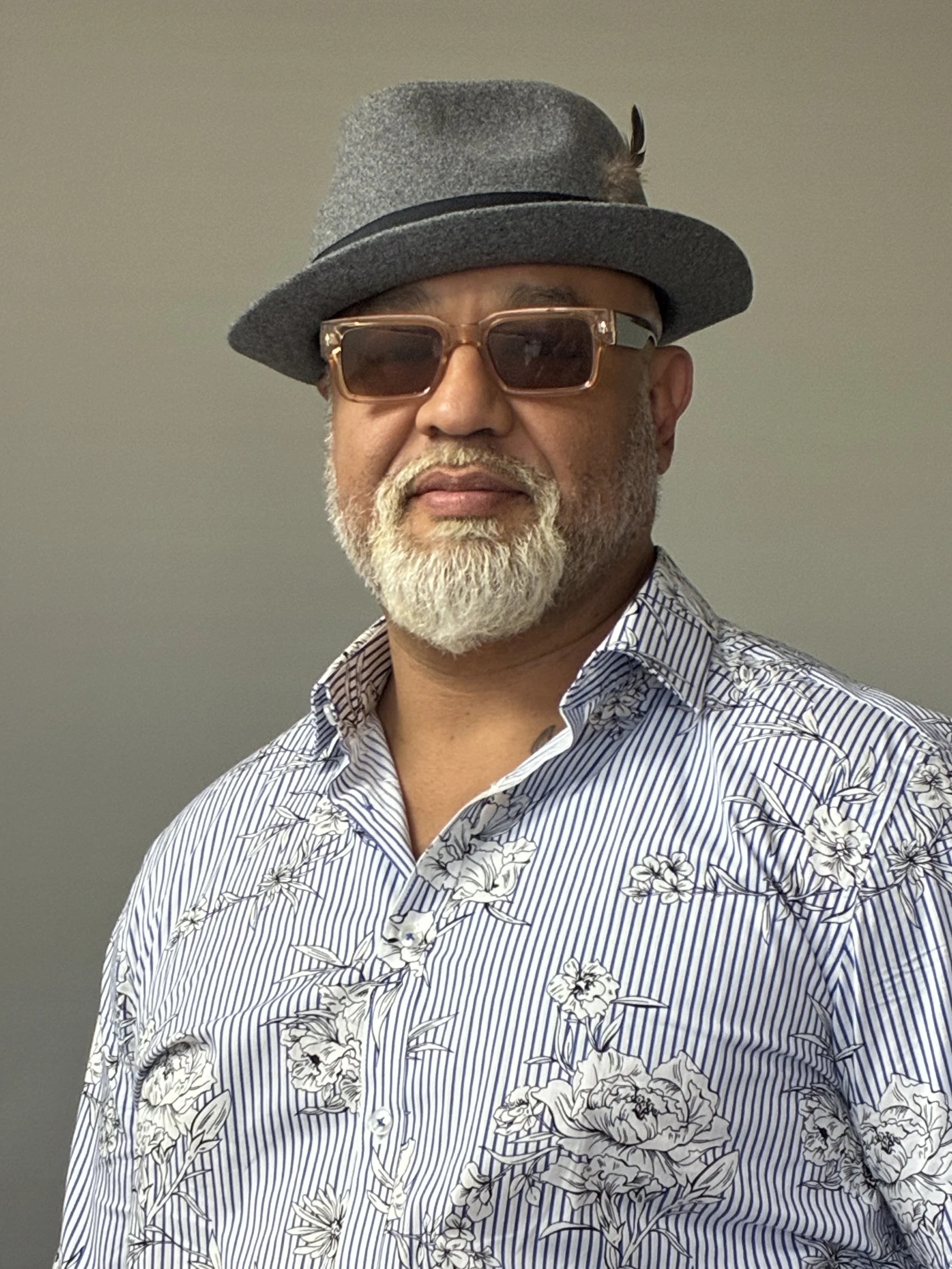 A middle-aged man with a white beard and mustache, wearing a gray fedora hat with a feather, sunglasses, and a blue and white striped floral shirt.