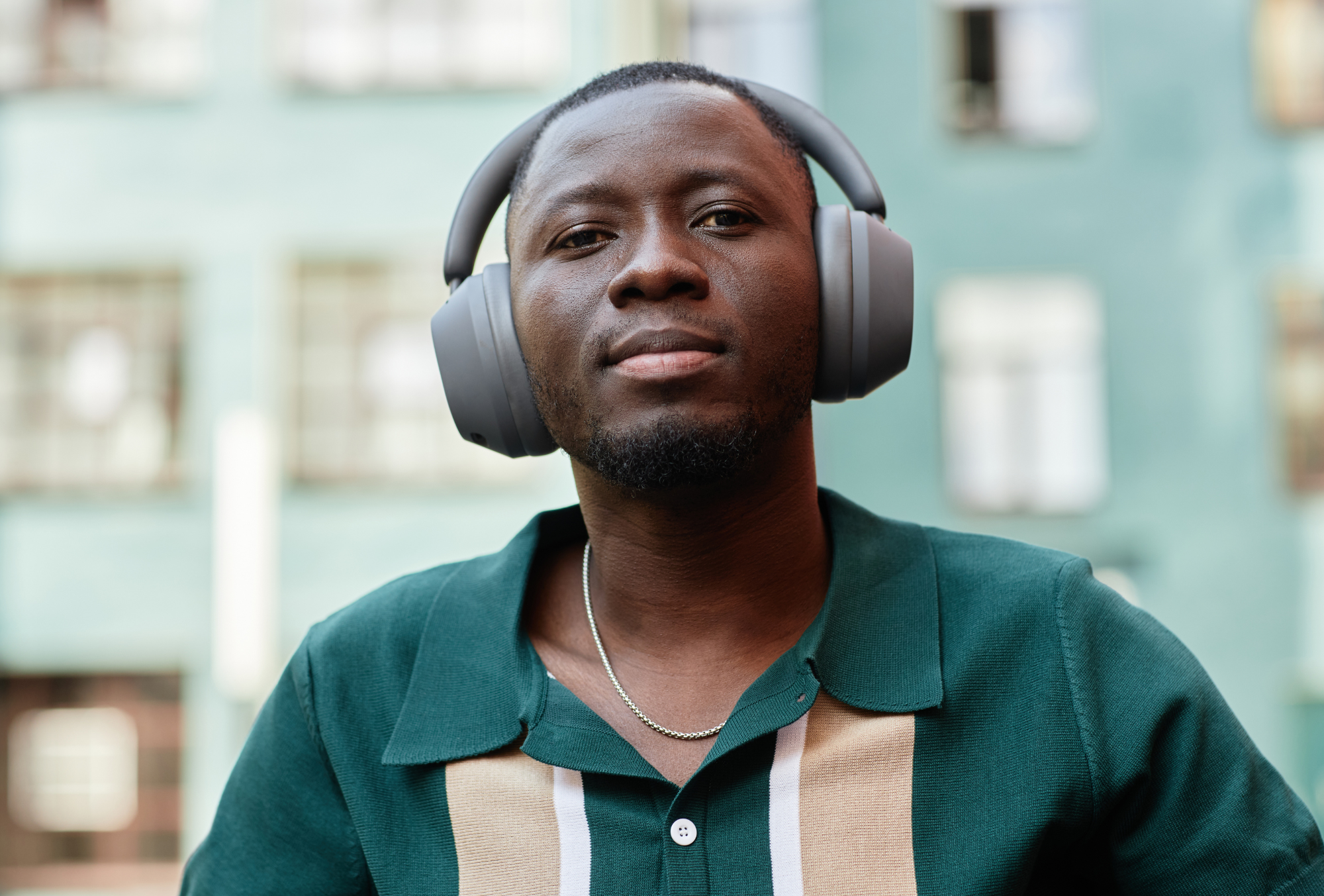 A man wearing a dark green shirt with beige and white accents and a silver chain necklace, standing outdoors with a building in the background. He is wearing large black over-ear headphones and looking confidently at the camera.
