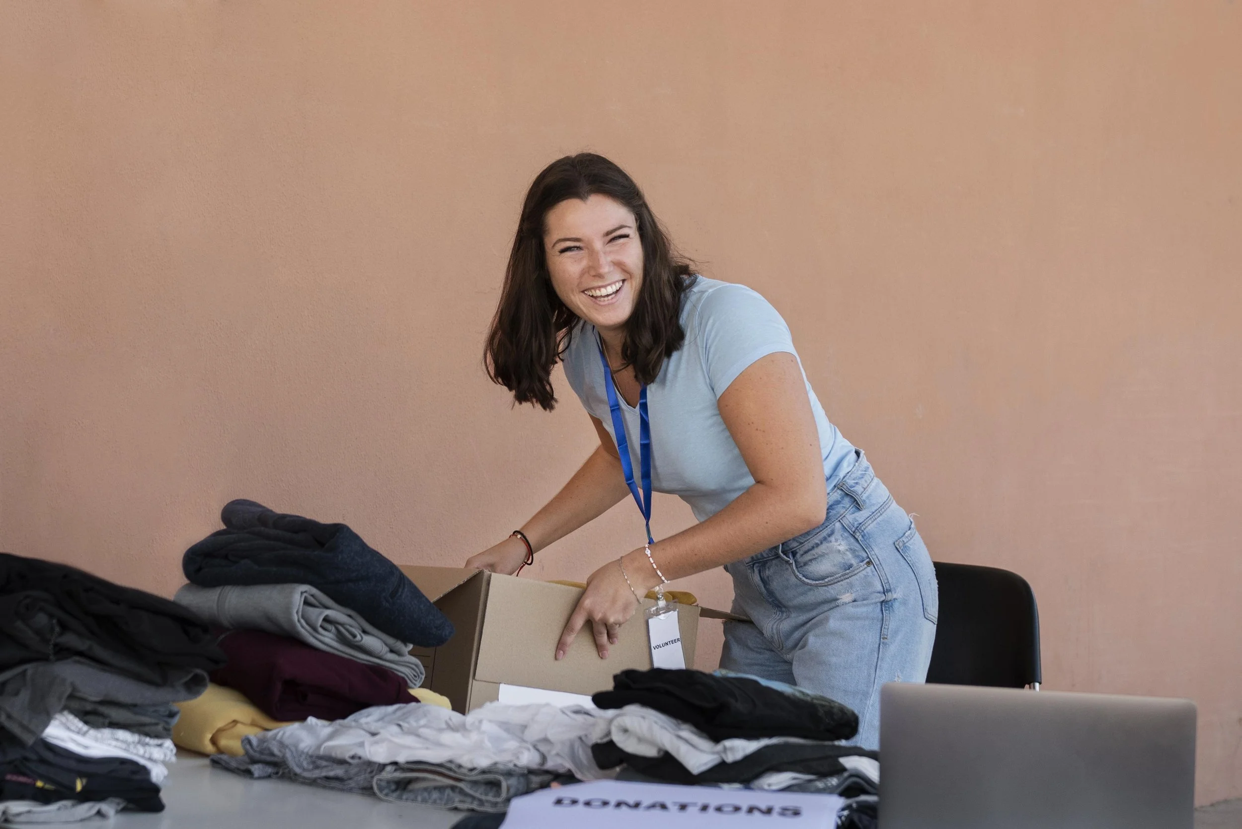 A smiling young woman with dark hair wearing a light blue t-shirt and jeans, organizing donated clothes on a table at a donation drive.