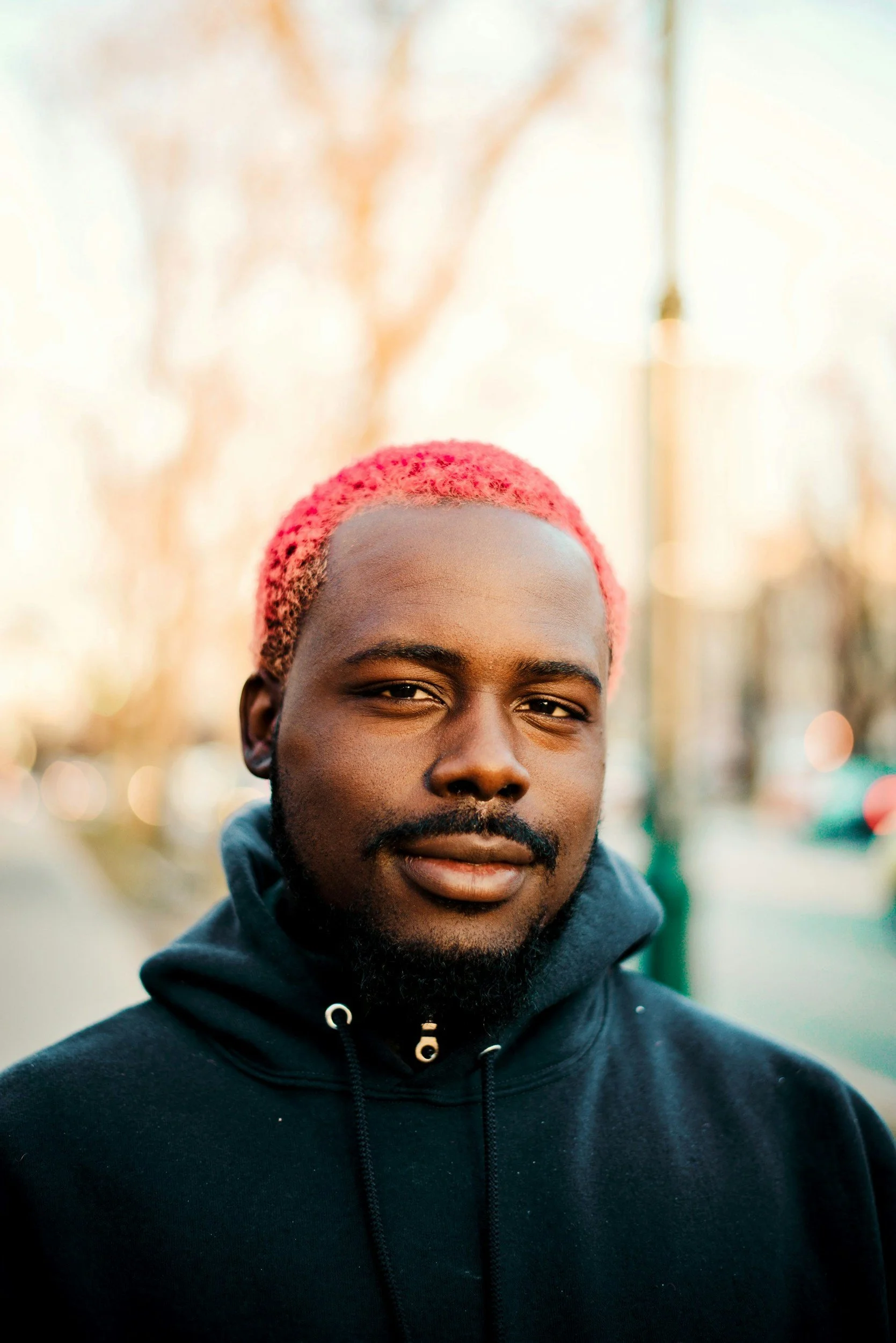 A close-up portrait of a young man with pink hair, a beard, and mustache, wearing a black hoodie, standing outdoors in a city environment with blurred trees and streetlights in the background.