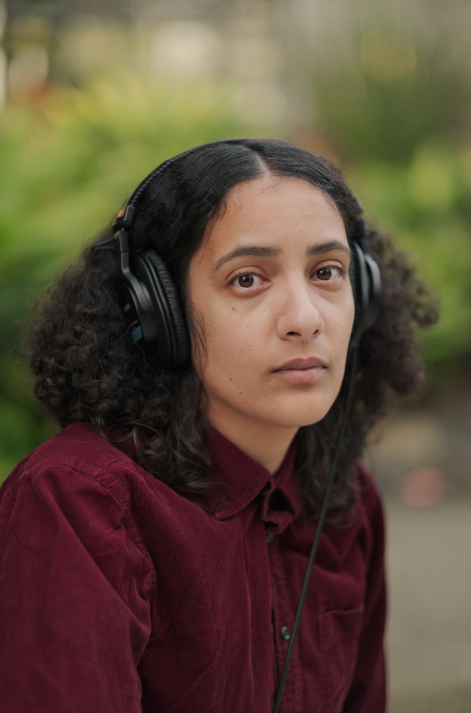 A young woman with curly dark hair wearing a maroon shirt and black headphones outdoors.