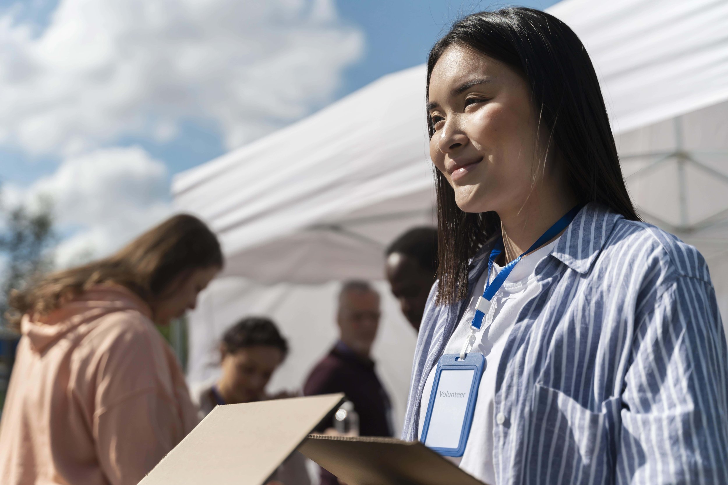 A woman wearing a Volunteer badge outdoors at a sunny event with multiple people and a white canopy in the background.