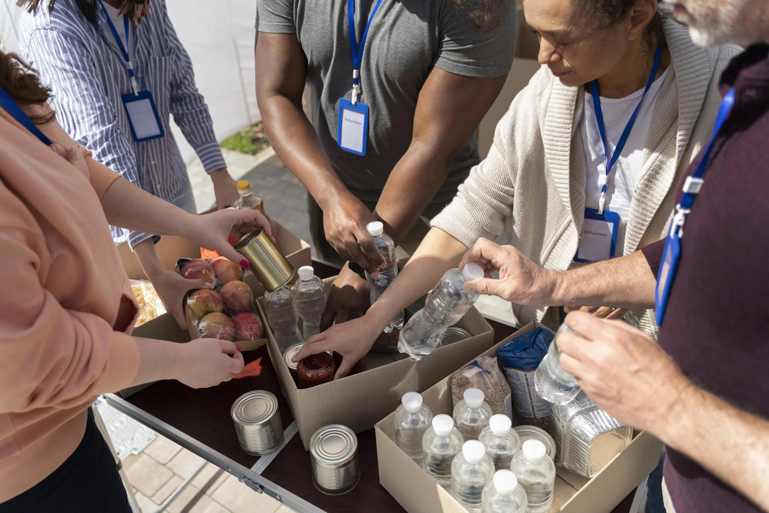 Volunteers are packing food and bottled water into boxes, with some canned goods and fresh apples visible, preparing supplies for distribution outdoors.