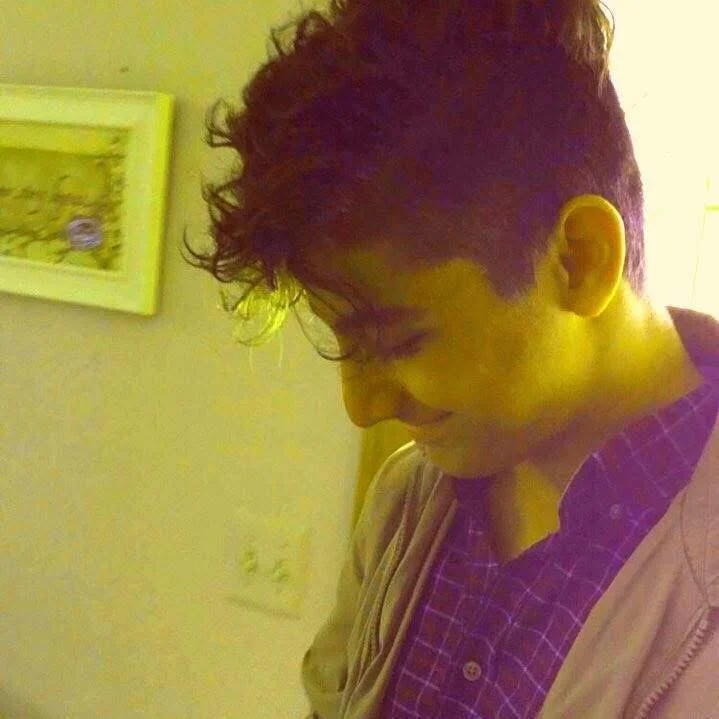 Danny Ashtiani smiling and looking downward, wearing a purple checkered shirt and beige jacket, standing indoors with a yellowish hue, with a framed picture on the wall behind him.