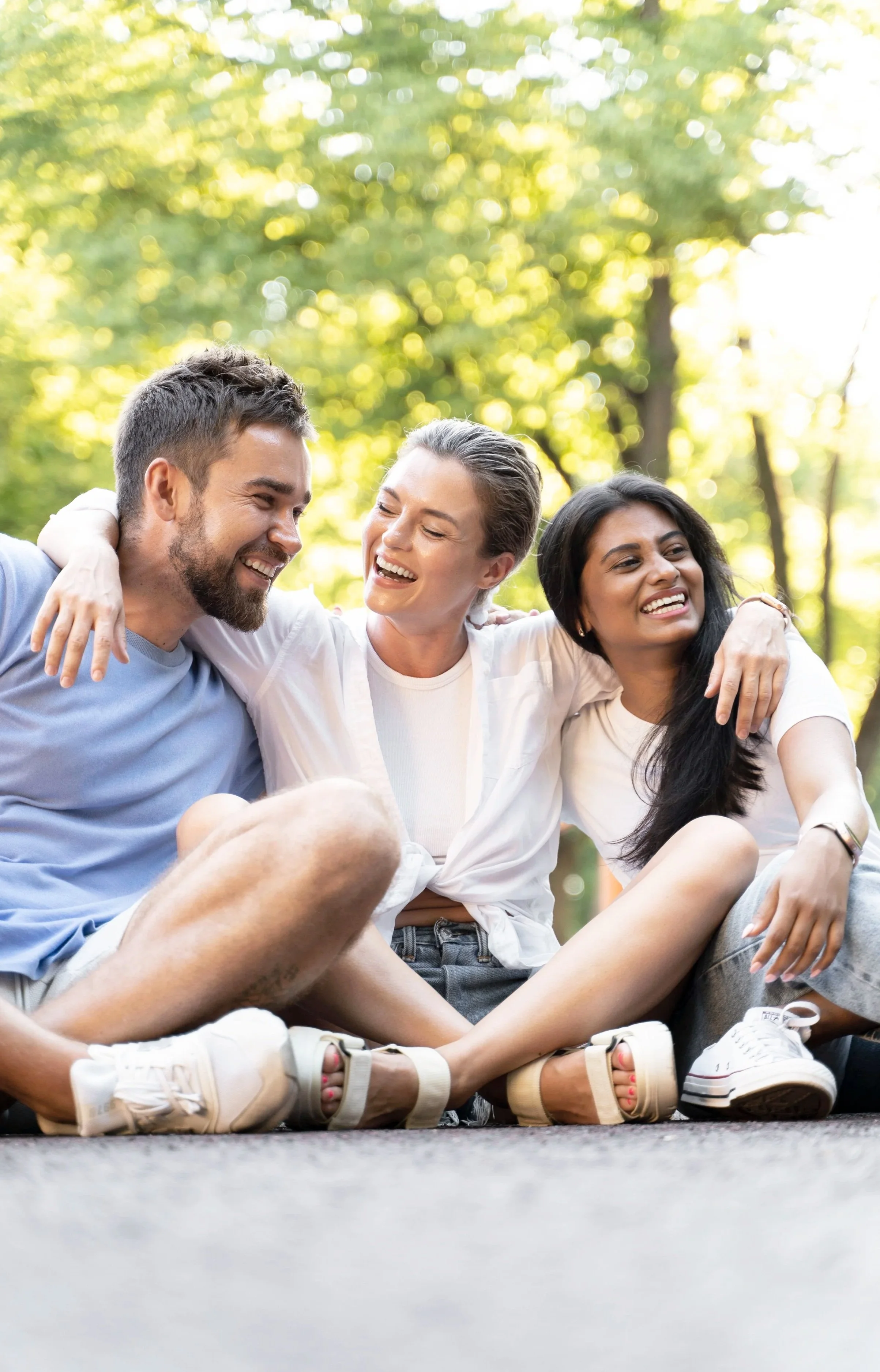 Three friends sitting on the ground outdoors, smiling and laughing together, surrounded by trees and sunlight.