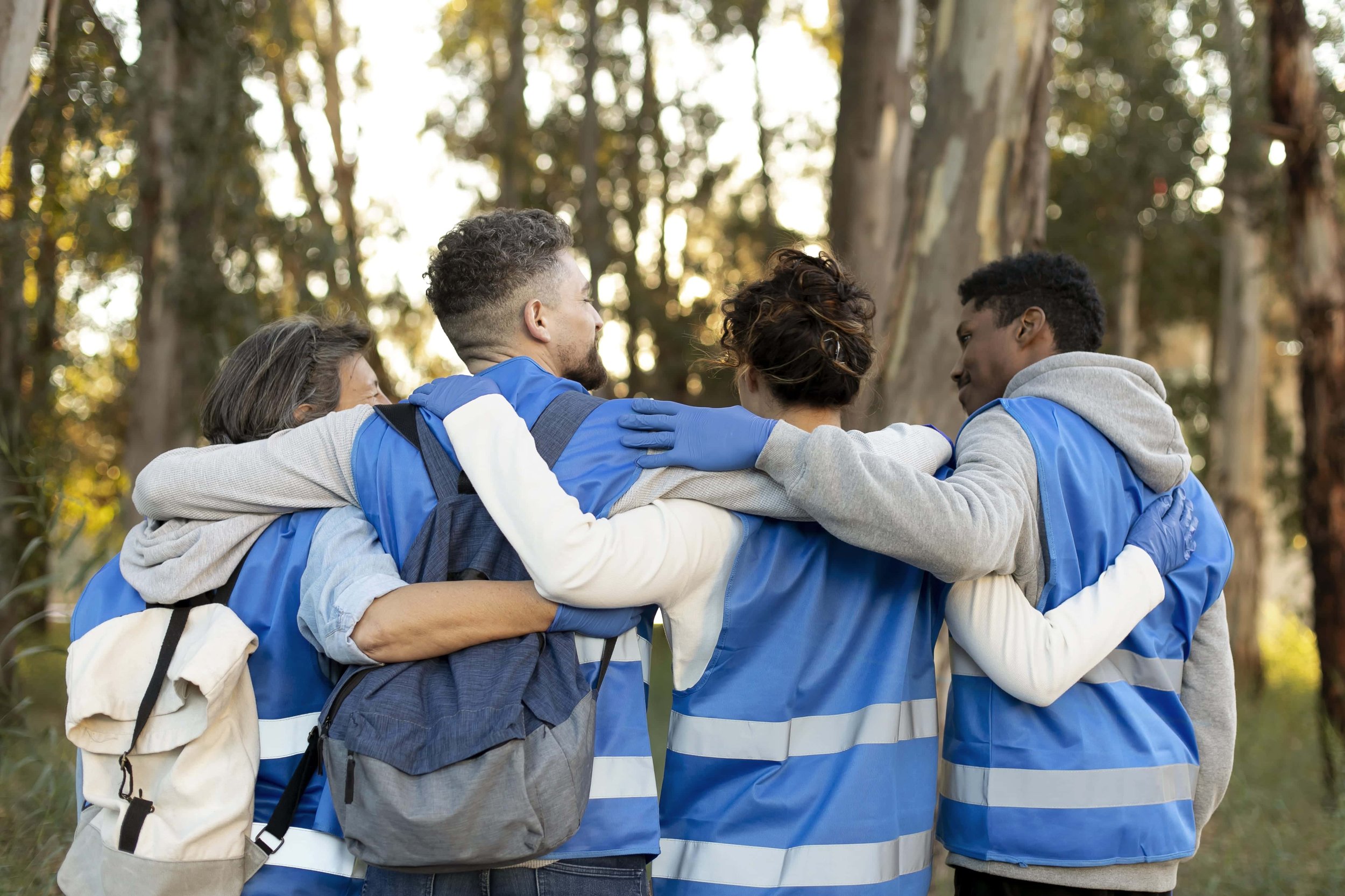 Group of five diverse volunteers in a forest, wearing blue safety vests, engaging in unity and teamwork by hugging each other.