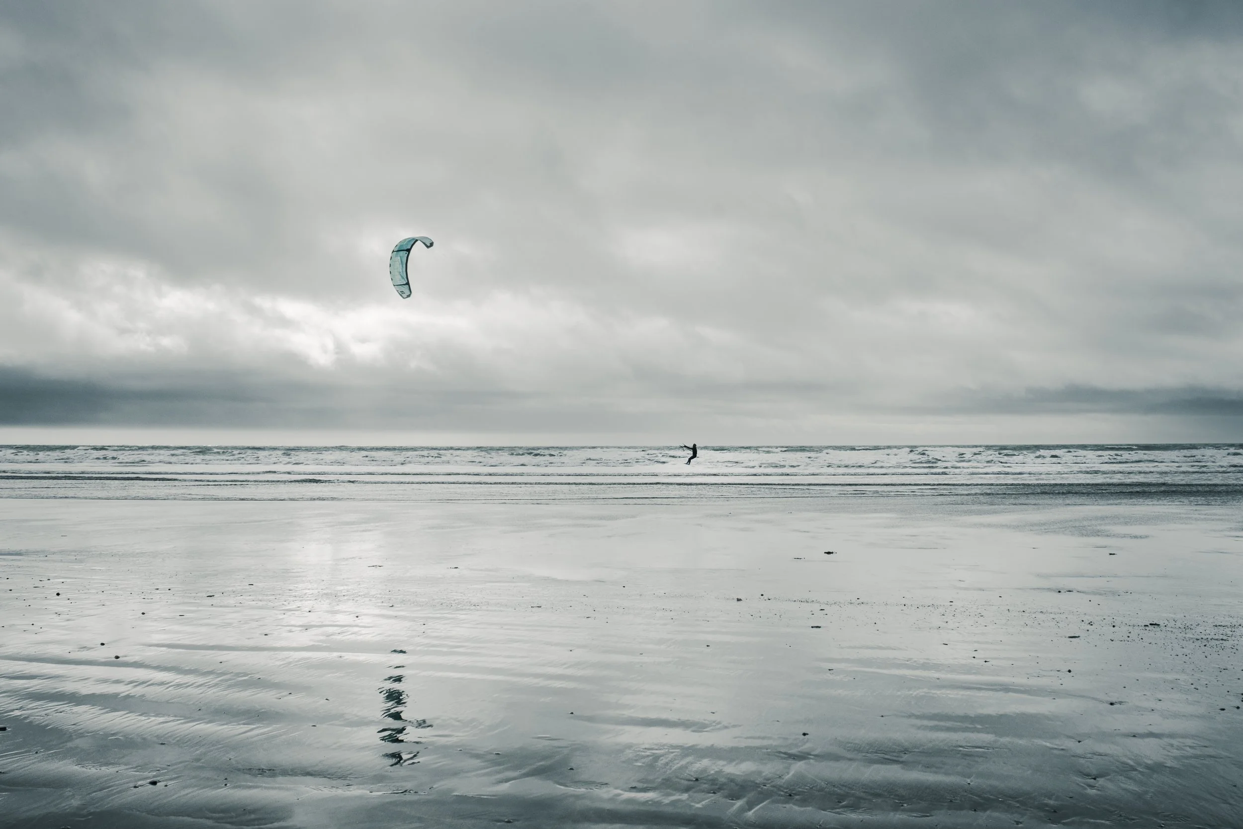 A person flying a kite or jumping on a wave on a cloudy beach with reflective wet sand.