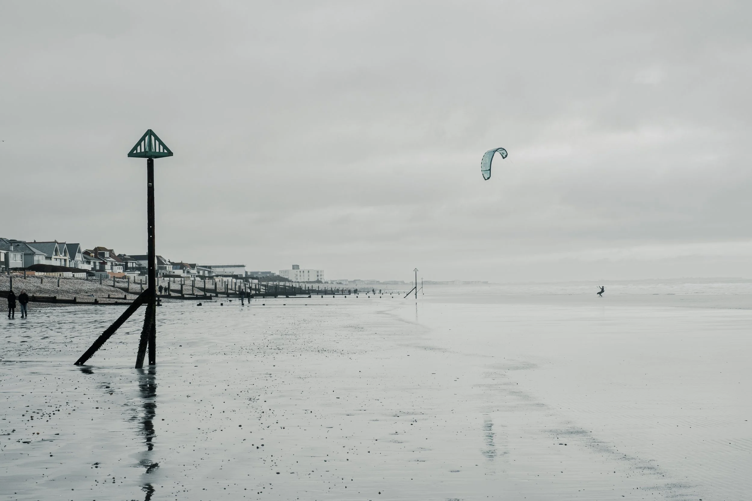 Overcast beach with a kite surfer, cloudy sky, houses along the shoreline, and waves in the background.