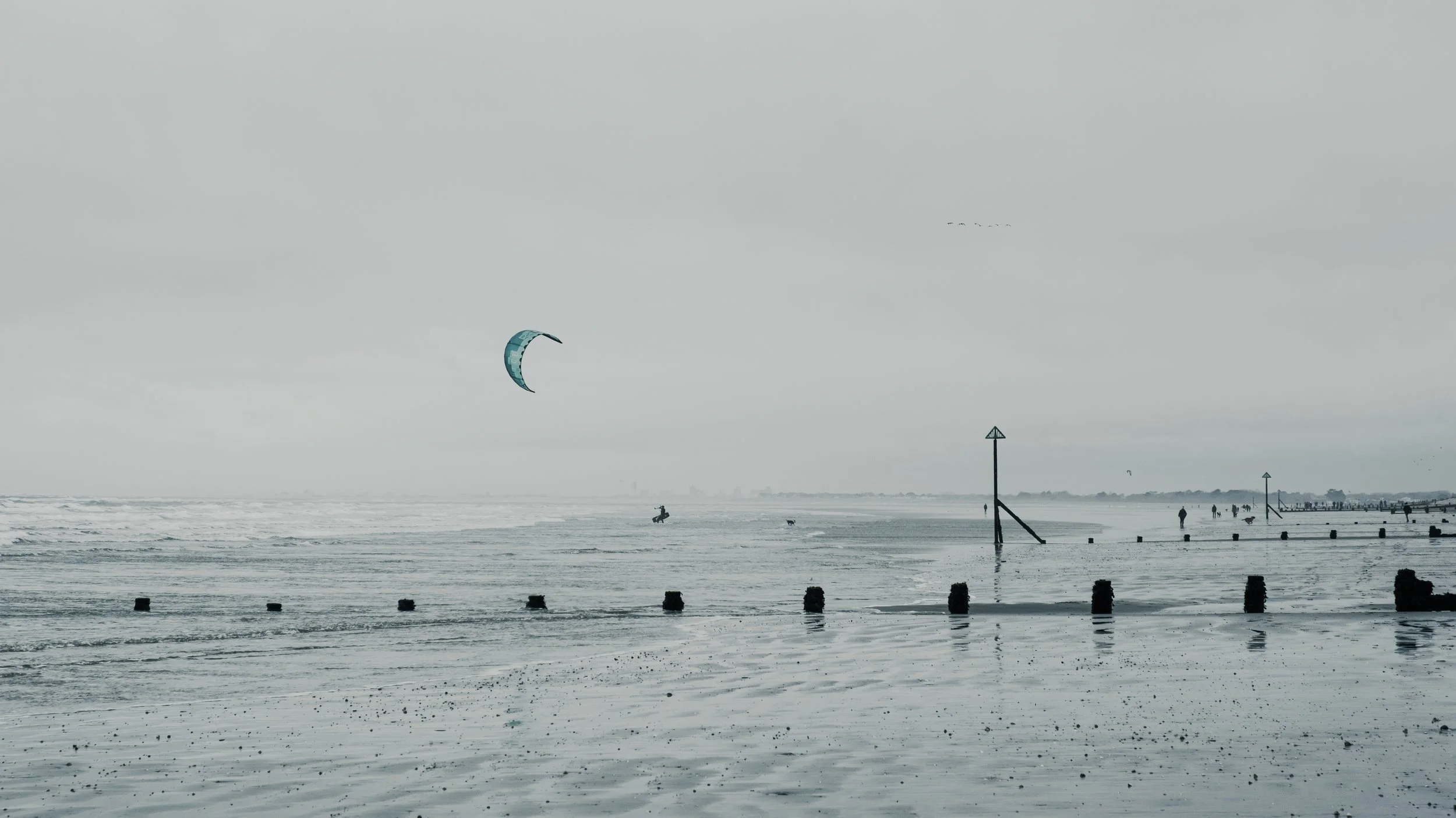 A cloudy beach scene with kite surfers, broken wooden posts, and distant shoreline.