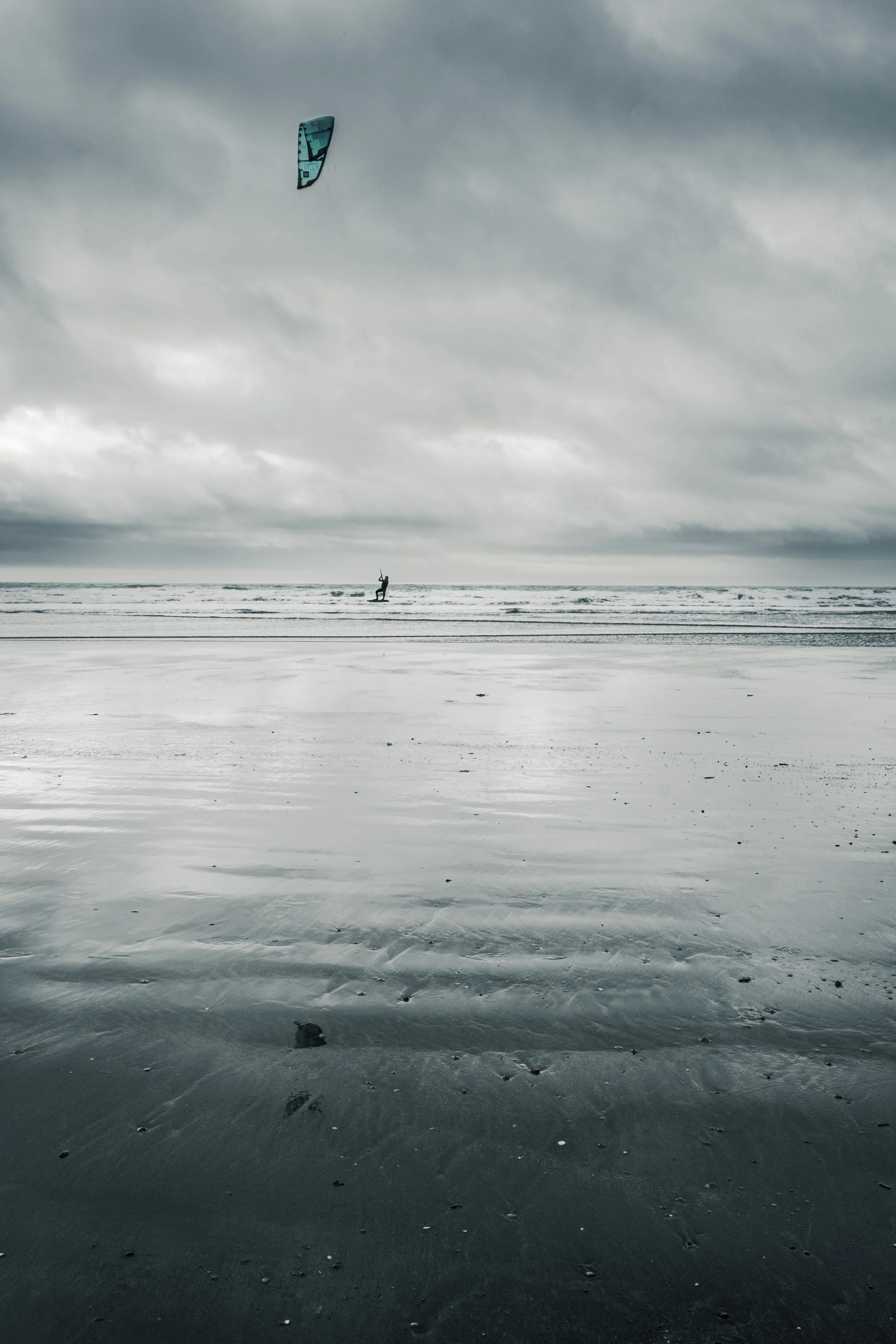 Person kiteboarding on the ocean under cloudy sky, with wet sandy beach in foreground.