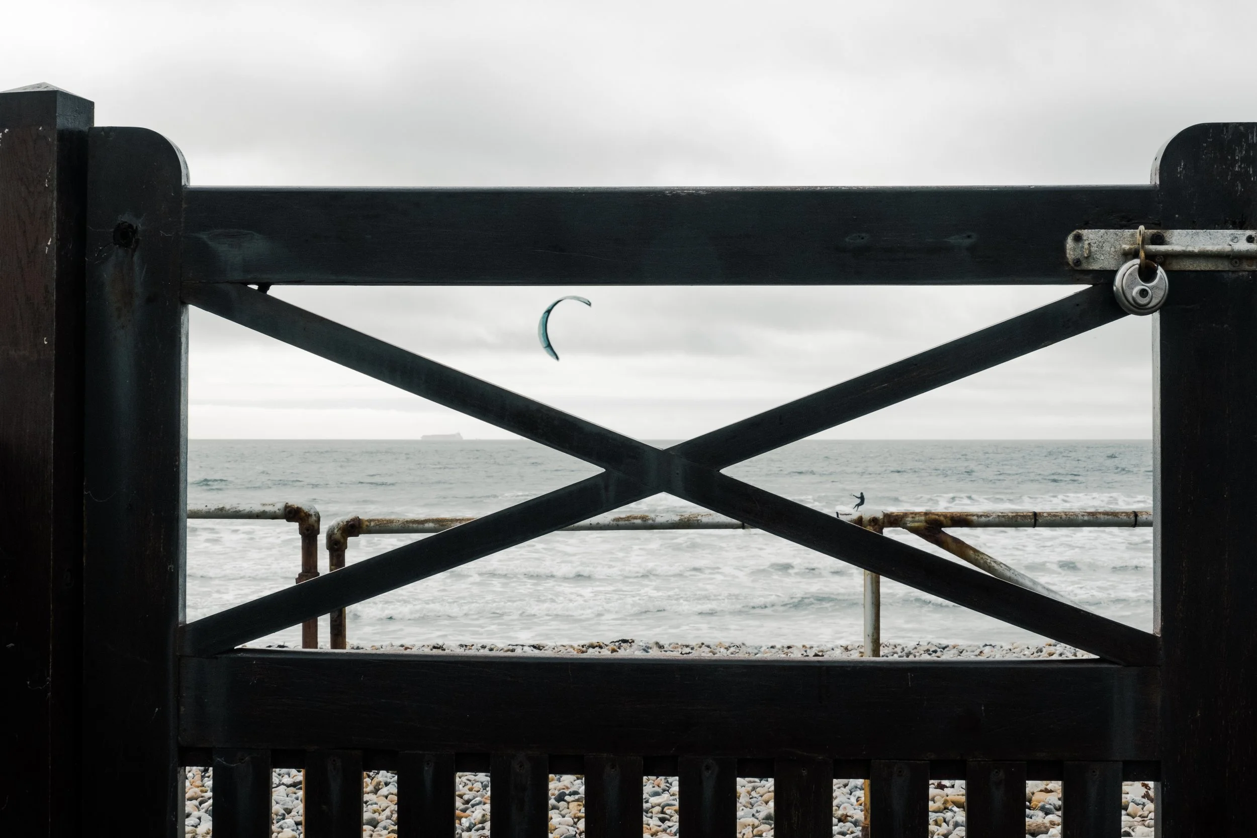 View of the ocean through a black wooden fence with a swirling padlock, a kite surfer in the water, and a ship in the distance on an overcast day.