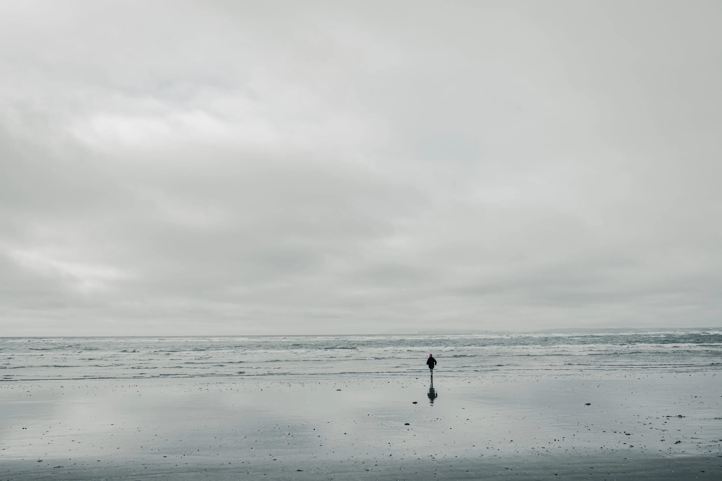 A solitary person walking on a wet beach under a cloudy gray sky, with the ocean in the background.
