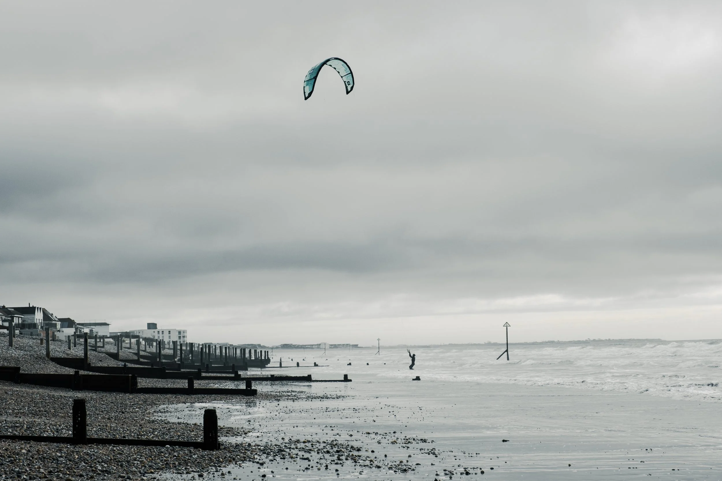 A person kite surfing on a cloudy day at the beach with houses on the shoreline and navigation markers in the water.