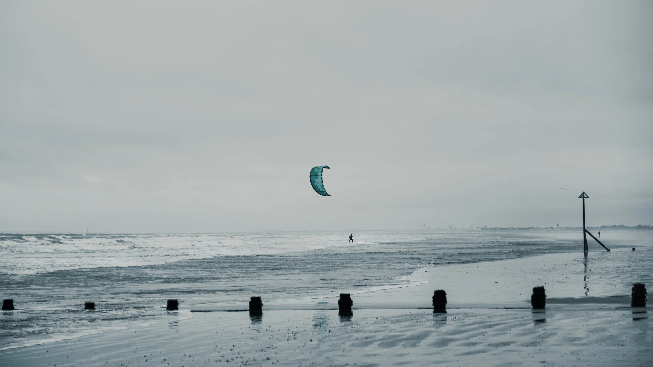 A person kiteboarding on a cloudy, overcast beach with choppy waters and a misty horizon.