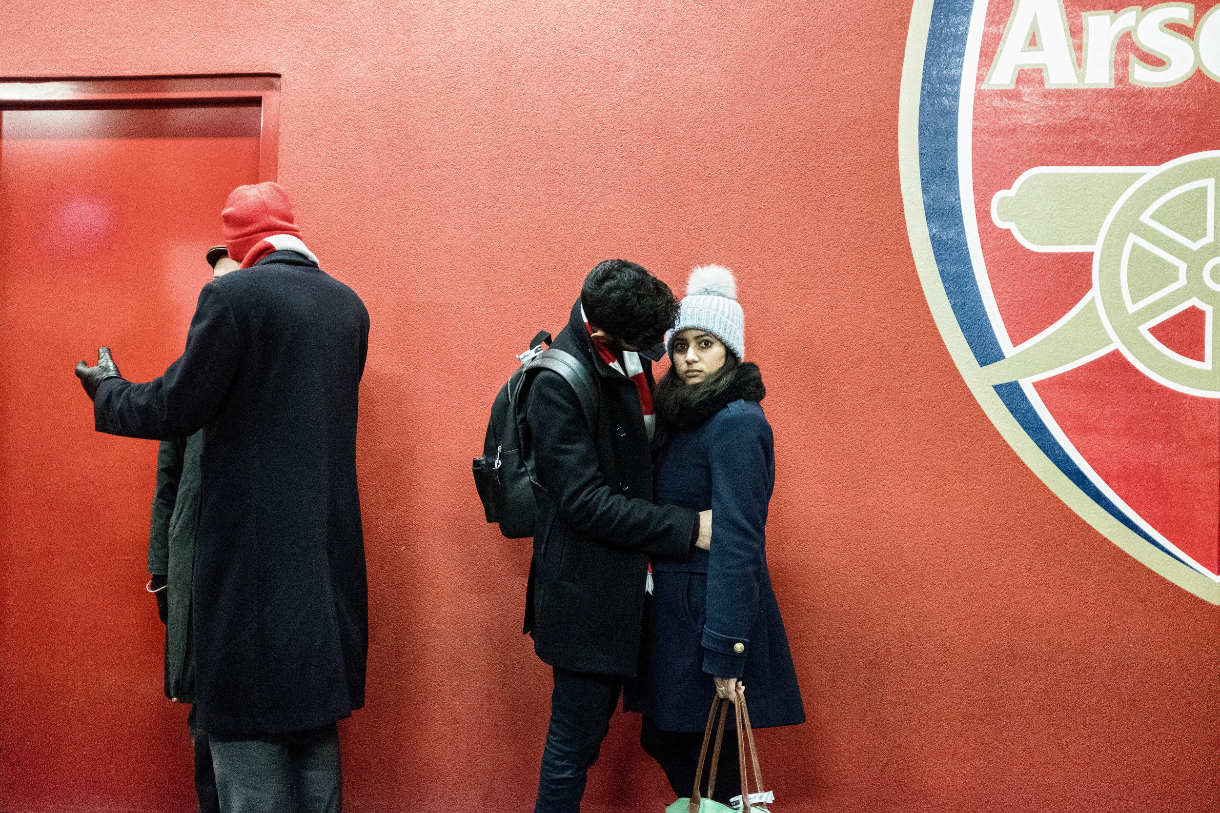 A group of three people standing against a red wall at a soccer stadium. Two are talking closely, and one is facing away from the camera, all bundled in winter clothing.