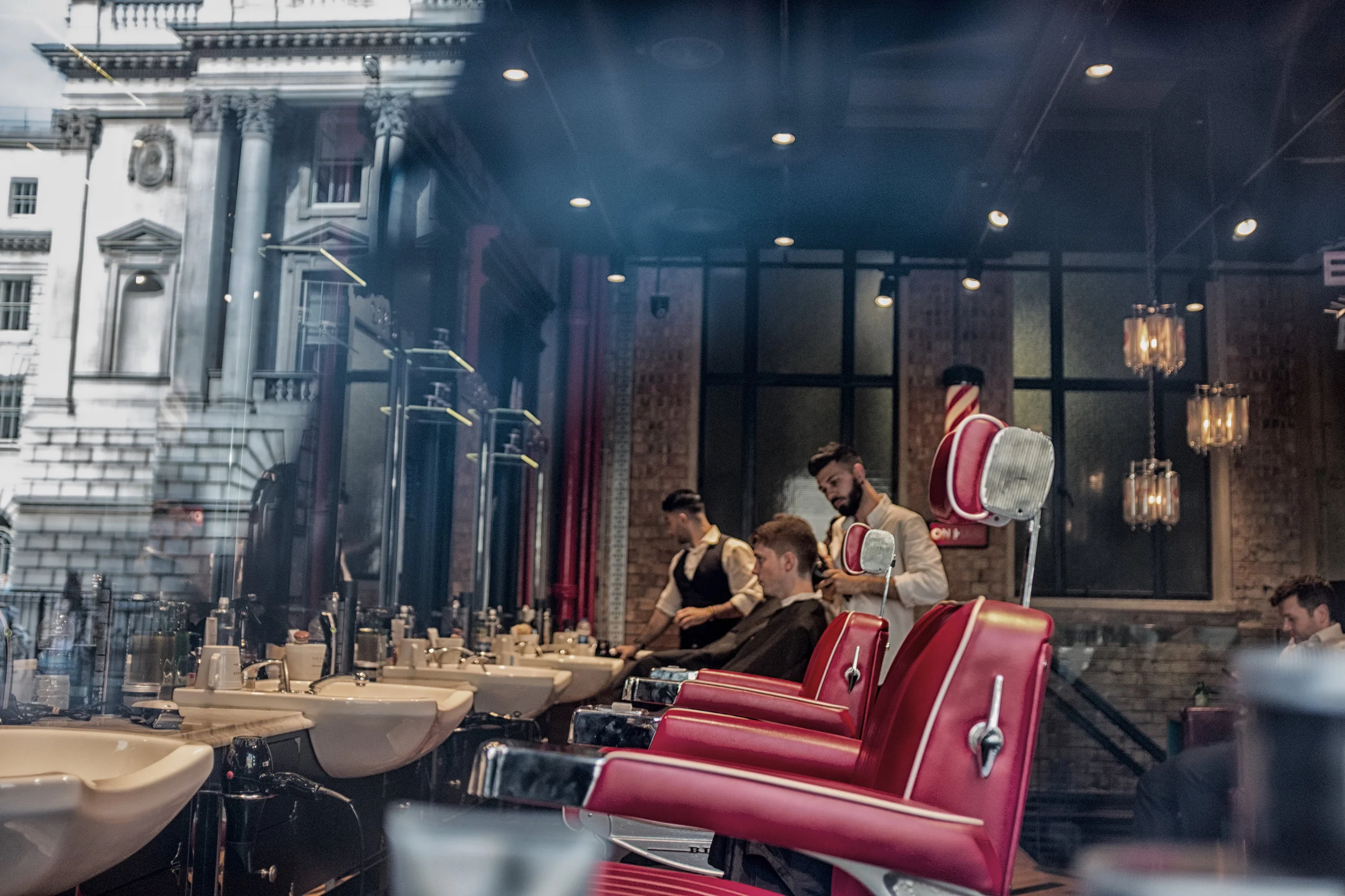 Interior of a modern barbershop with red chairs, mirrors, and sinks, with barbers attending to a client, while a glass window reflects the street outside.
