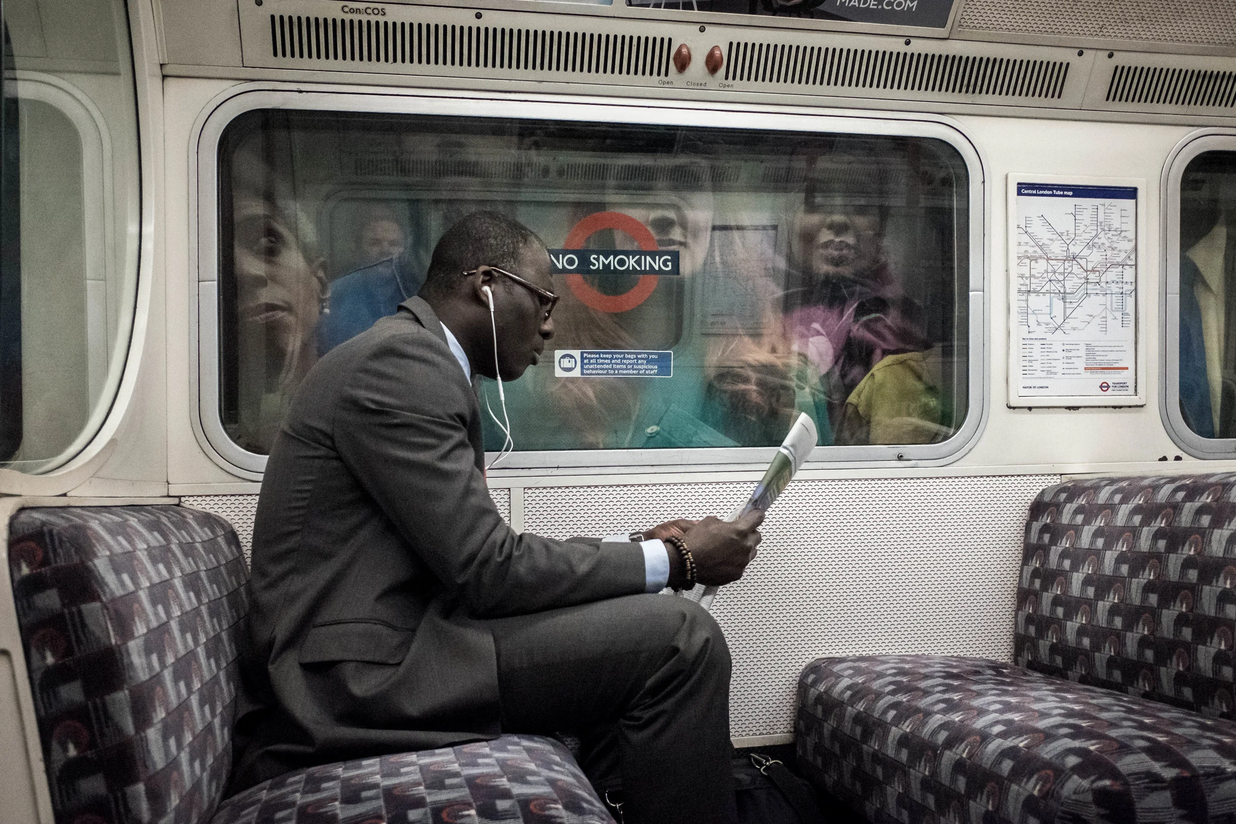 A man in a suit sitting on a train, wearing glasses and earphones, reading a newspaper while others look out the window behind the glass.