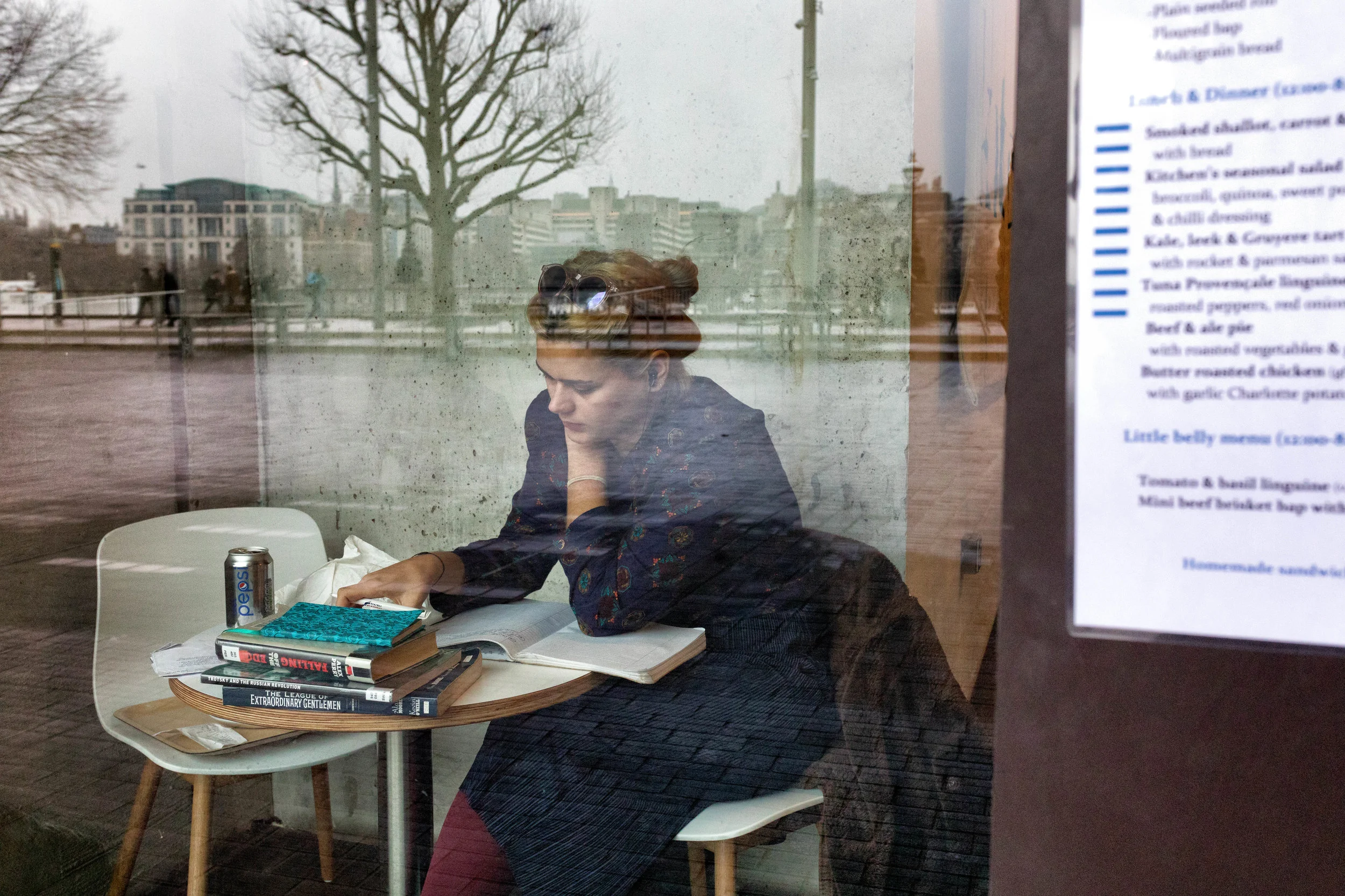 A woman with red hair and sunglasses on her head, sitting at a small round table inside a cafe, seen through a glass window. She is reading a book, with a can of Pepsi, a stack of books, and an open notebook on the table. The window reflects an outdo