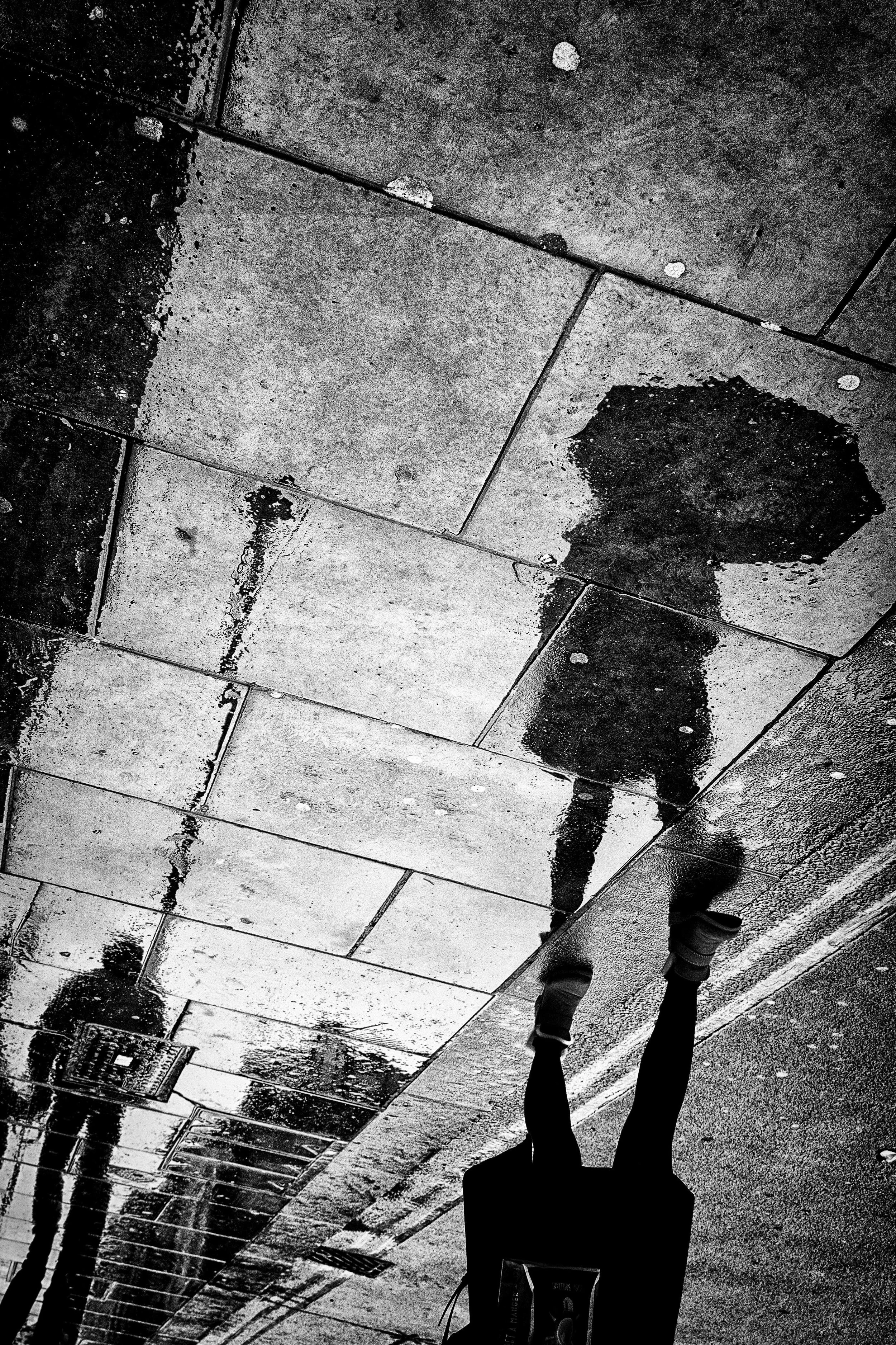 Black and white photo of a street sidewalk with wet patches, a person walking away, and their reflection visible in a puddle.