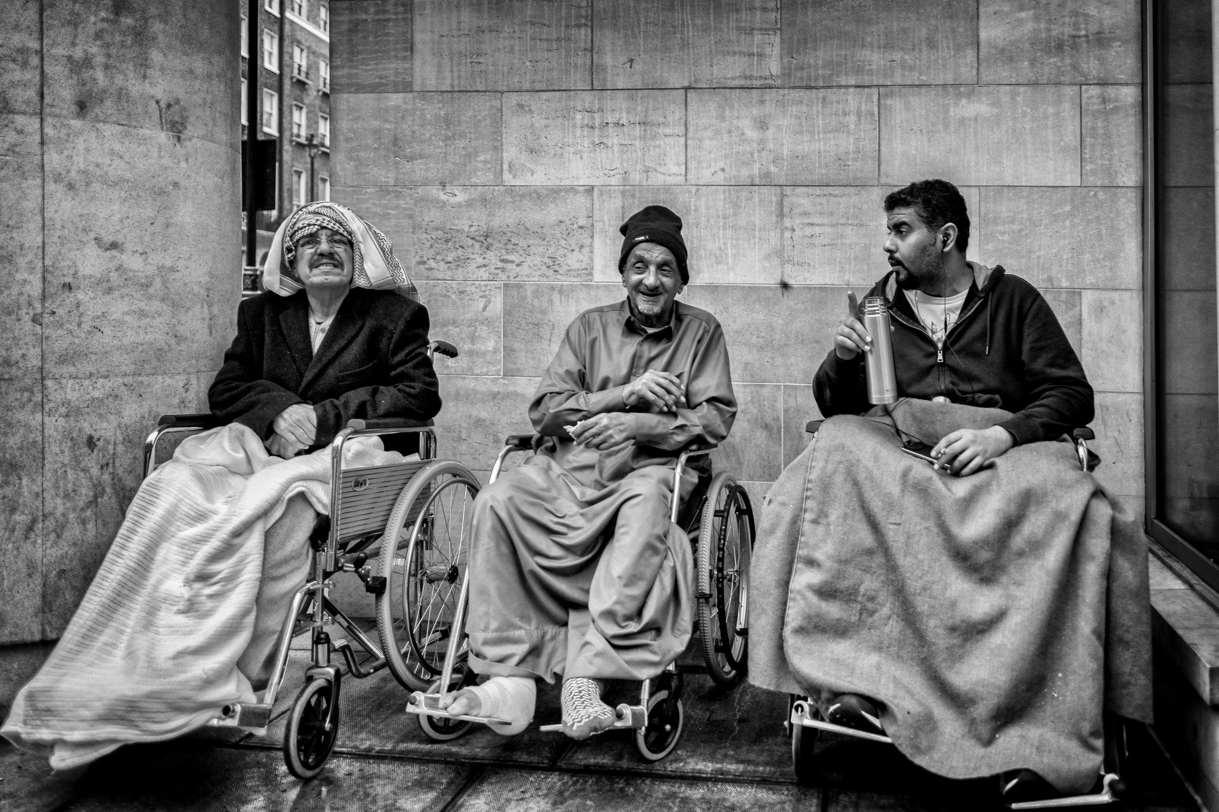 Three people sitting on a city sidewalk: an elderly man in a wheelchair with a headscarf, a man in a wheelchair with a beanie, and a young man in a wheelchair with a beverage bottle, all engaged in conversation.
